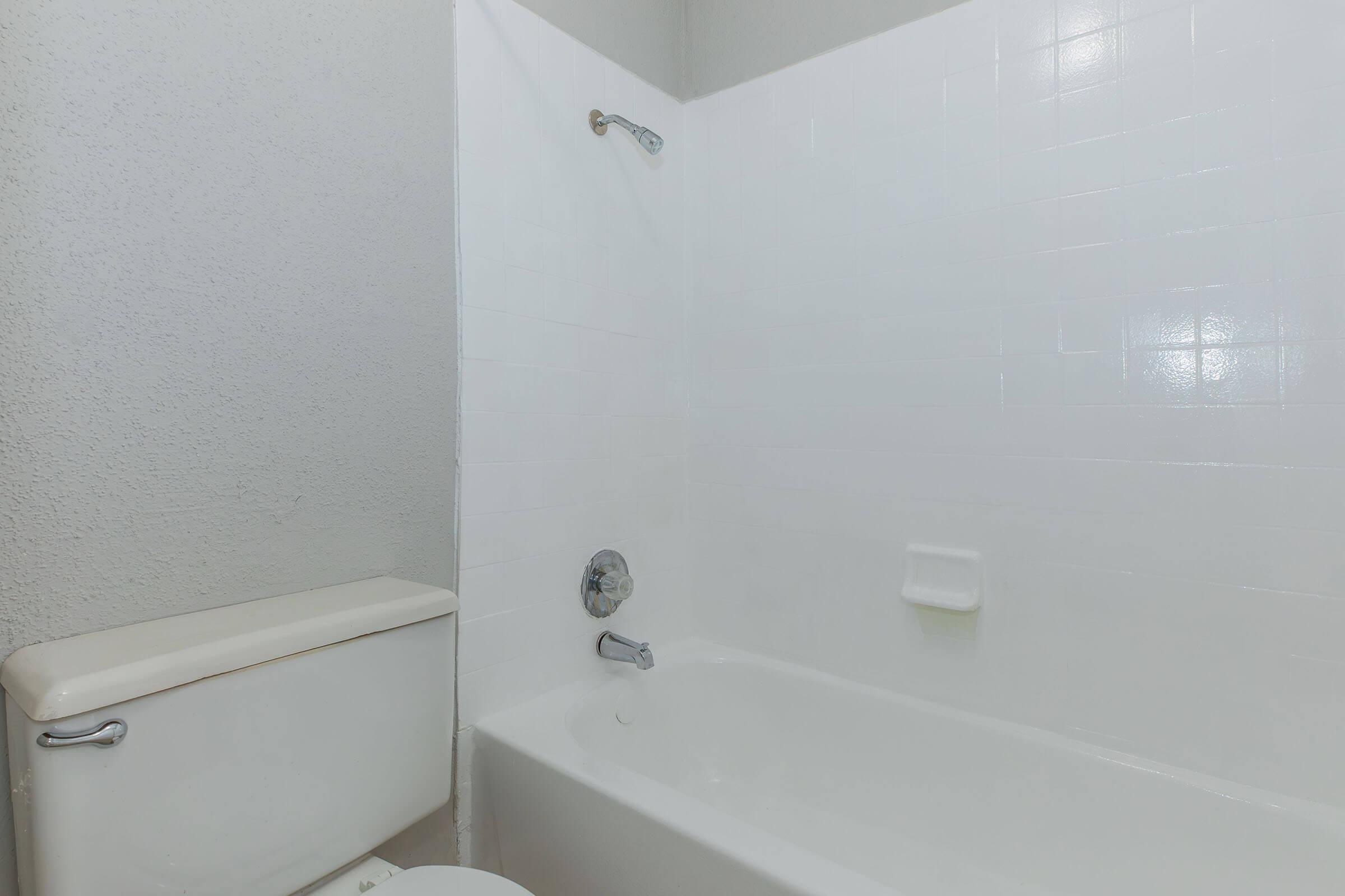 A clean, modern bathroom featuring a white bathtub and shower combination. The wall is tiled with glossy white tiles, and there is a small soap holder on the wall. A toilet is visible to the left, complementing the overall minimalistic design. The space appears bright and well-maintained.