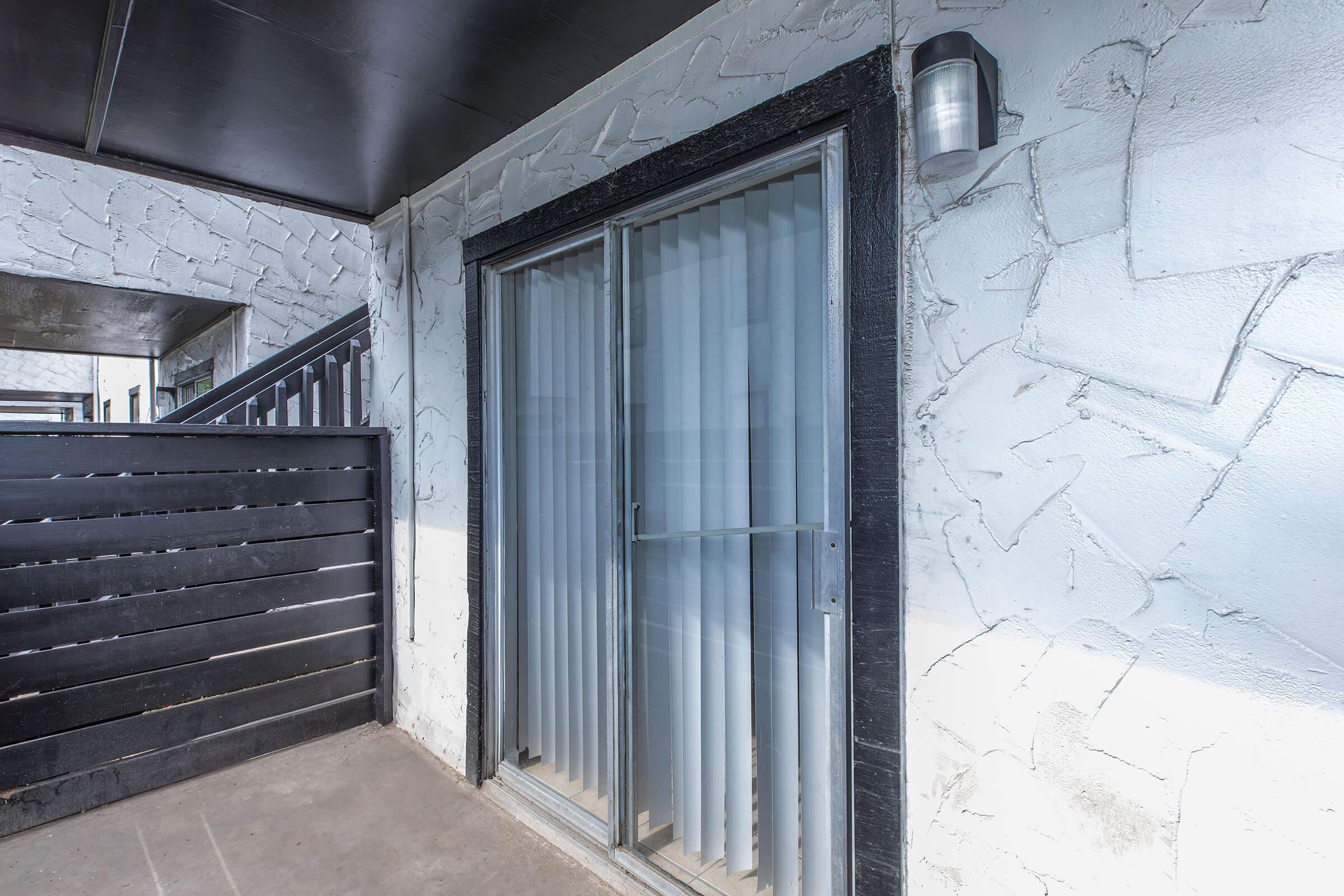 A modern sliding glass door with vertical blinds, set in a textured light-colored wall. There is a black wooden fence to the left and a wall-mounted light fixture beside the door. The entrance appears to be part of a contemporary building exterior.