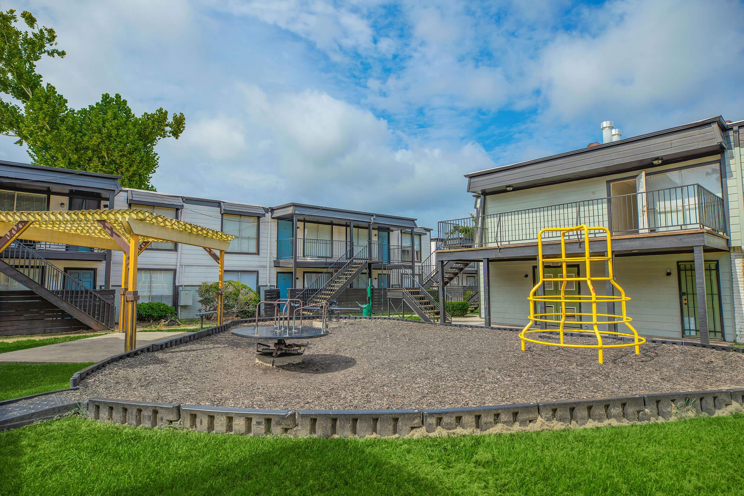 Playground area featuring a yellow climbing structure, a spinning merry-go-round, and grassy surroundings. Two multi-level apartment buildings with balconies are visible in the background under a partly cloudy sky. A wooden gazebo is also present, creating a welcoming outdoor space for residents.