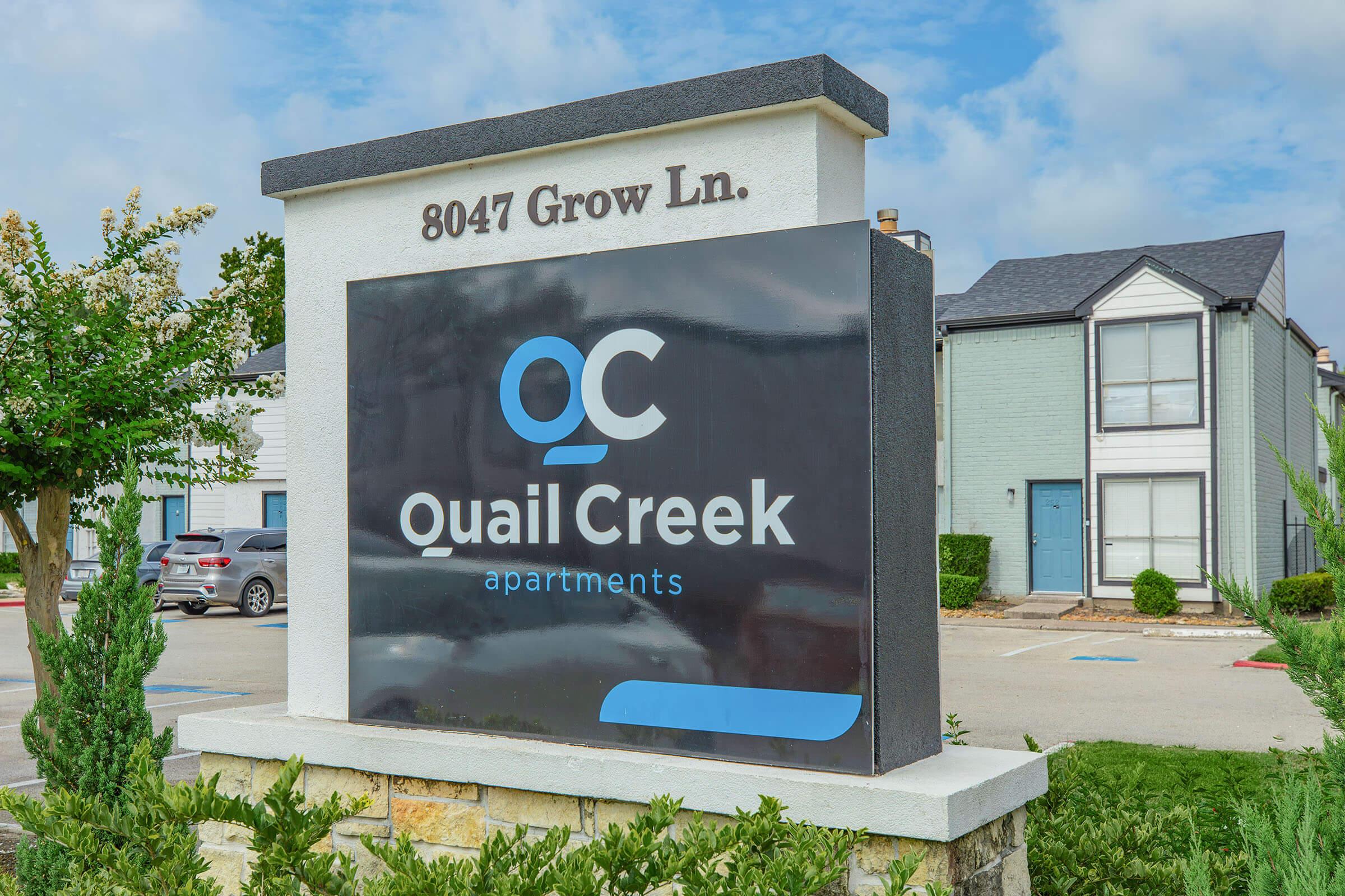 Sign for Quail Creek Apartments located at 8047 Grow Ln. The sign features the logo with "QC" and includes the name "Quail Creek apartments." The background shows a cloudy sky and landscaped area with parking spaces and apartment buildings visible.
