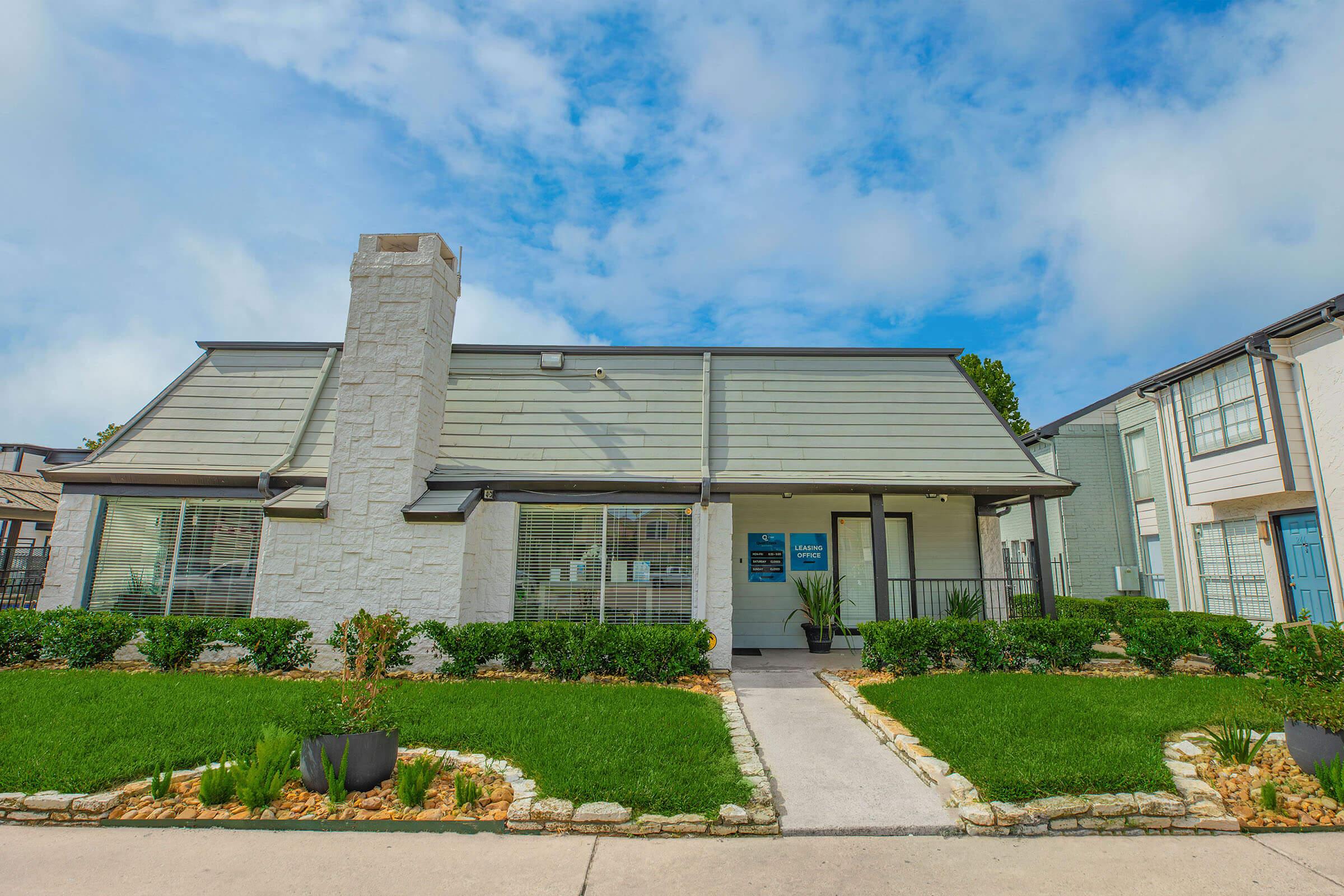 A well-maintained modern house featuring a stone exterior and a prominent chimney. The front yard is landscaped with lush green grass and decorative stones, framed by low bushes. The entrance includes large windows and a welcoming porch, with a sign visible on the building. The sky above is partly cloudy.