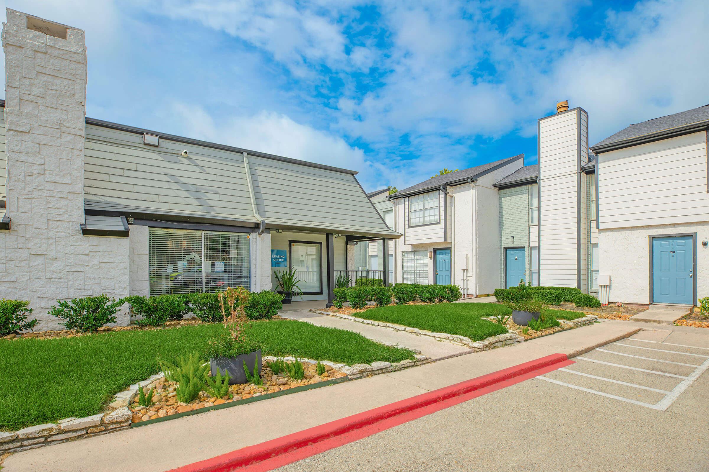 A well-maintained multi-unit apartment complex with a landscaped garden, featuring green grass, small bushes, and stone pathways. The building has a modern design with a mix of gray siding and white accents, under a partly cloudy blue sky. Parking spaces are visible in the foreground.