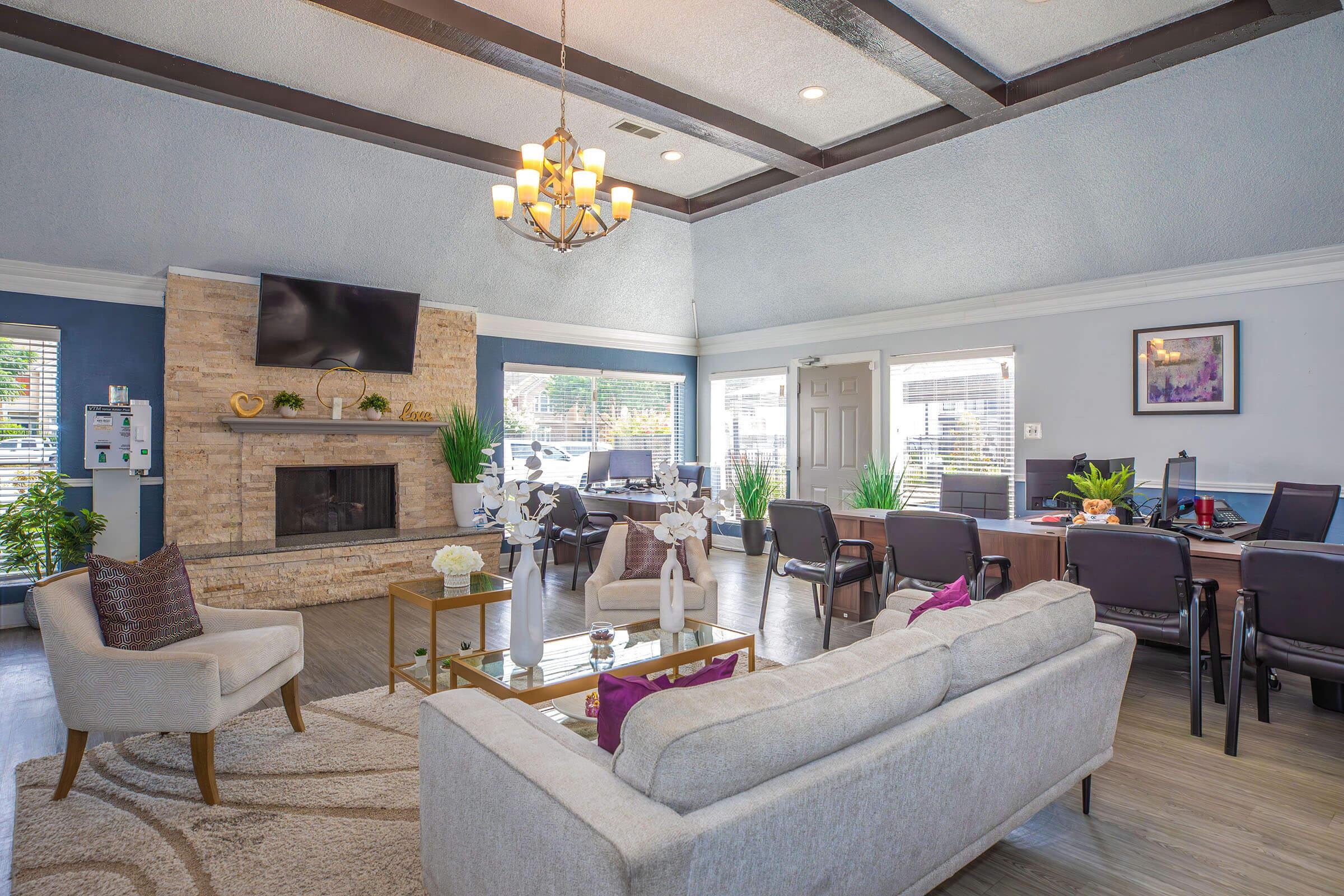 A modern living room with a cozy seating area featuring a light-colored couch, decorative pillows, and a glass coffee table. There is a stone fireplace with a television above it, and large windows letting in natural light. The space includes plants and a dining area with chairs and a table, creating a welcoming atmosphere.