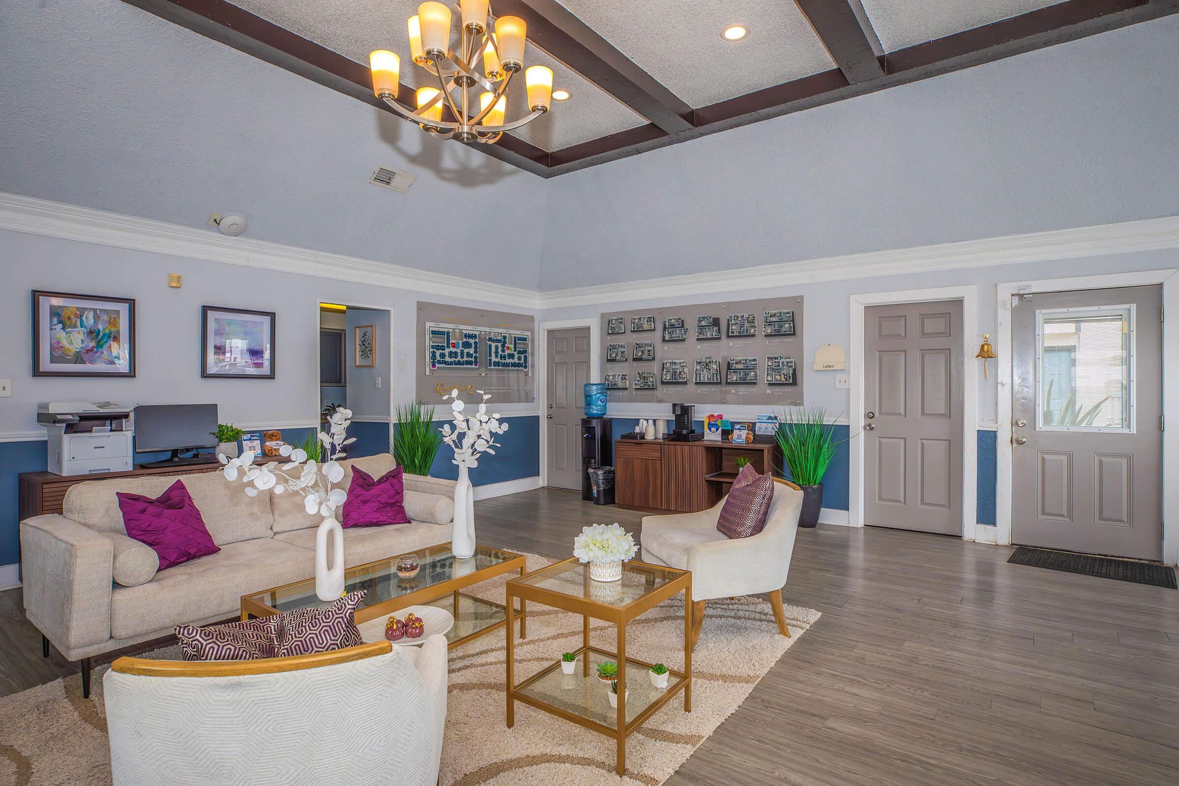A modern living room with a high ceiling, featuring a beige sofa with purple cushions, a glass coffee table, and decorative plants. The room has a stylish chandelier, framed artwork on the walls, a desk area, and a door leading outside, creating a welcoming and cozy atmosphere.