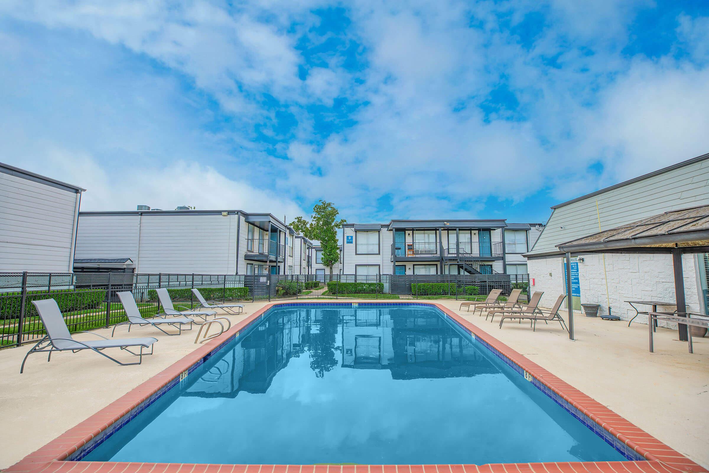 A clear blue swimming pool surrounded by lounge chairs, set in a residential area with modern apartment buildings in the background. The sky is partly cloudy, creating a serene atmosphere for relaxation and leisure.