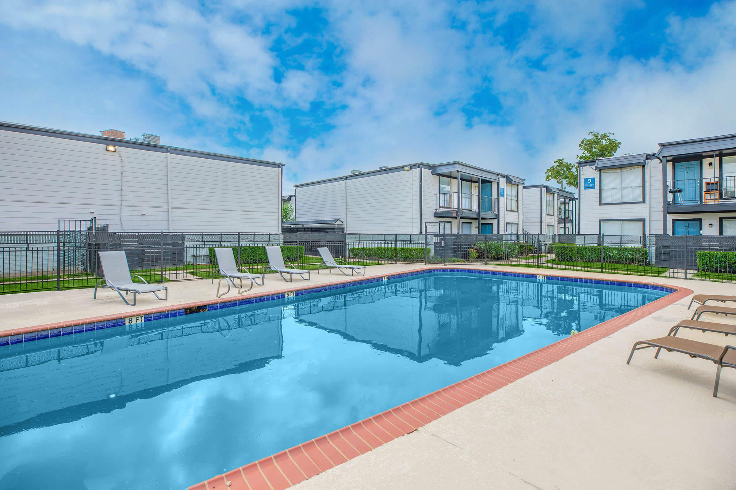 A refreshing outdoor swimming pool surrounded by lounge chairs, with modern apartment buildings in the background. The pool is clear and inviting, framed by a well-maintained patio area and green landscaping under a bright blue sky with some clouds.