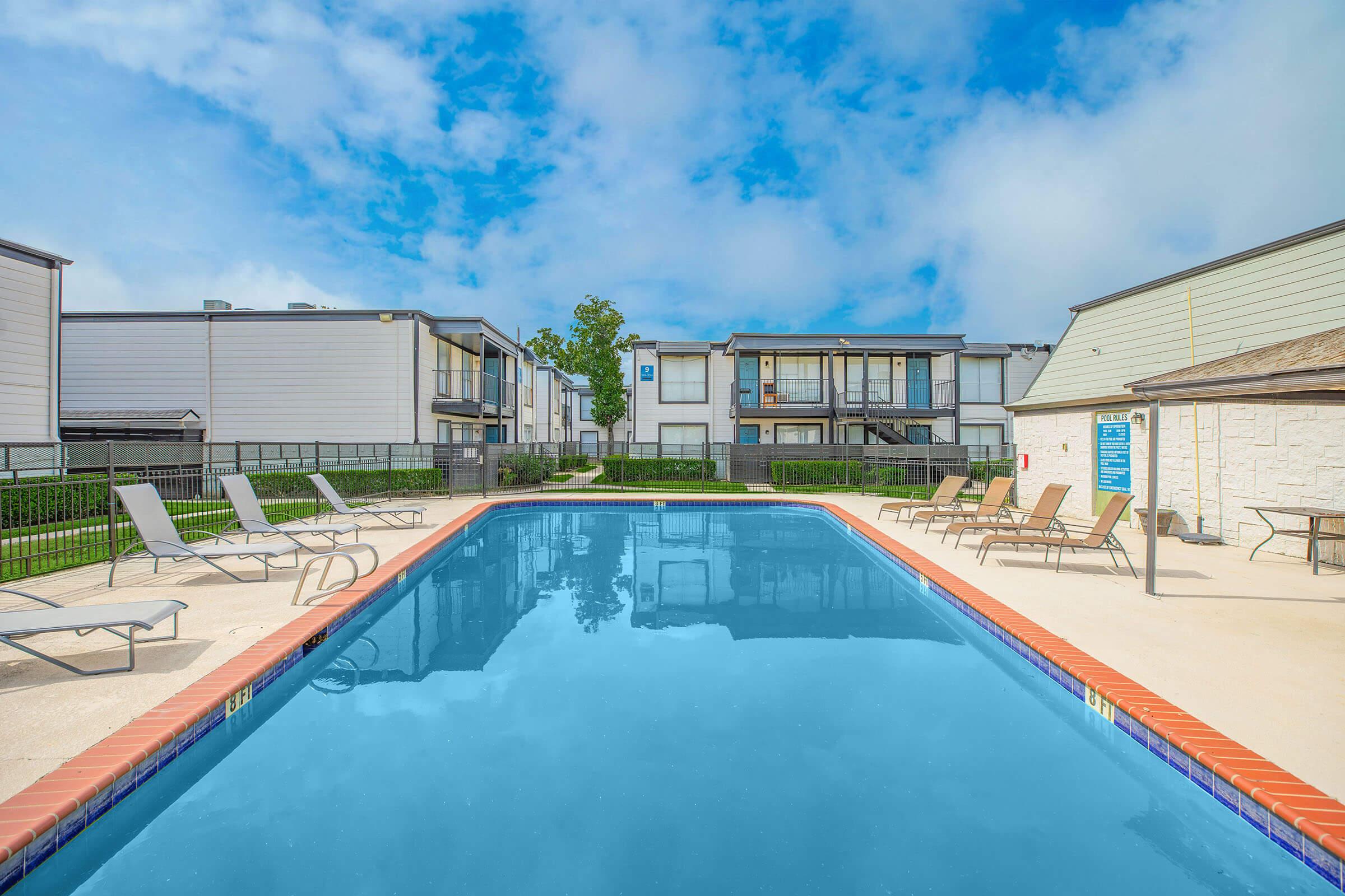 A swimming pool surrounded by lounge chairs, with apartment buildings in the background. The sky is partly cloudy, creating a serene atmosphere. The pool area is fenced, providing privacy for residents enjoying the outdoor space.
