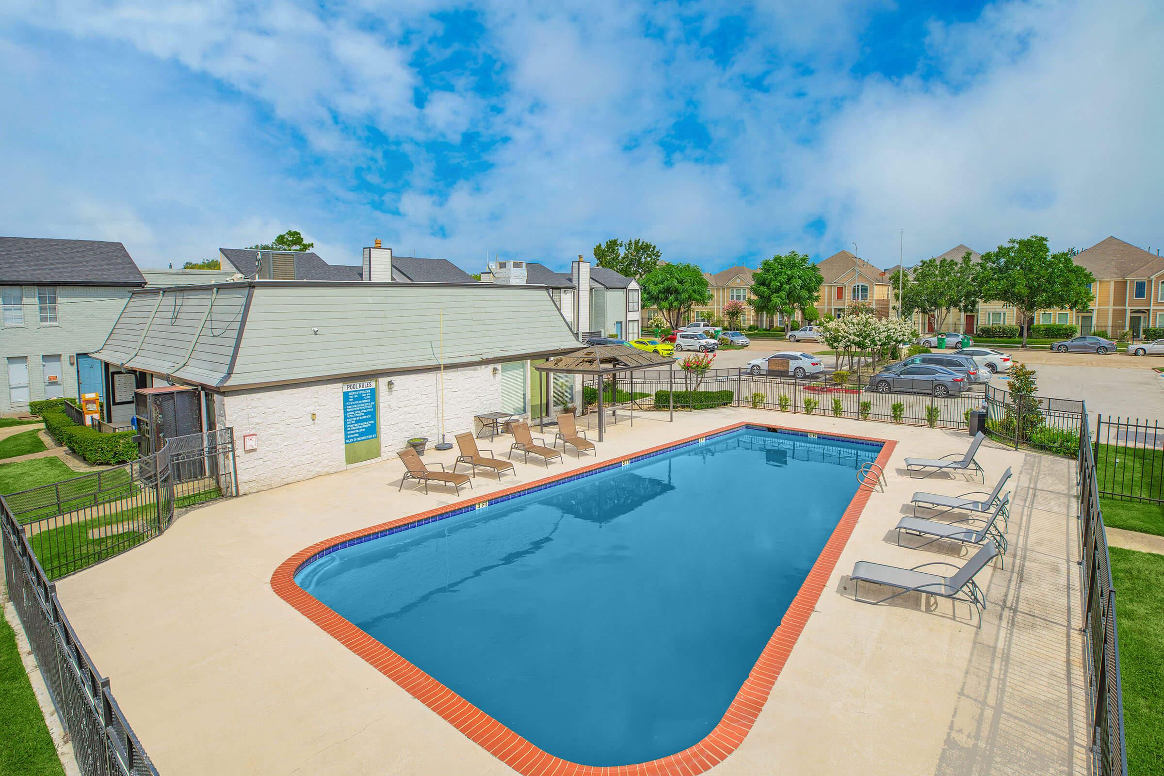 A rectangular swimming pool surrounded by a concrete deck and lounge chairs. A small gazebo is nearby, and the scene includes green grass and well-maintained landscaping. Residential buildings are visible in the background under a partly cloudy sky.