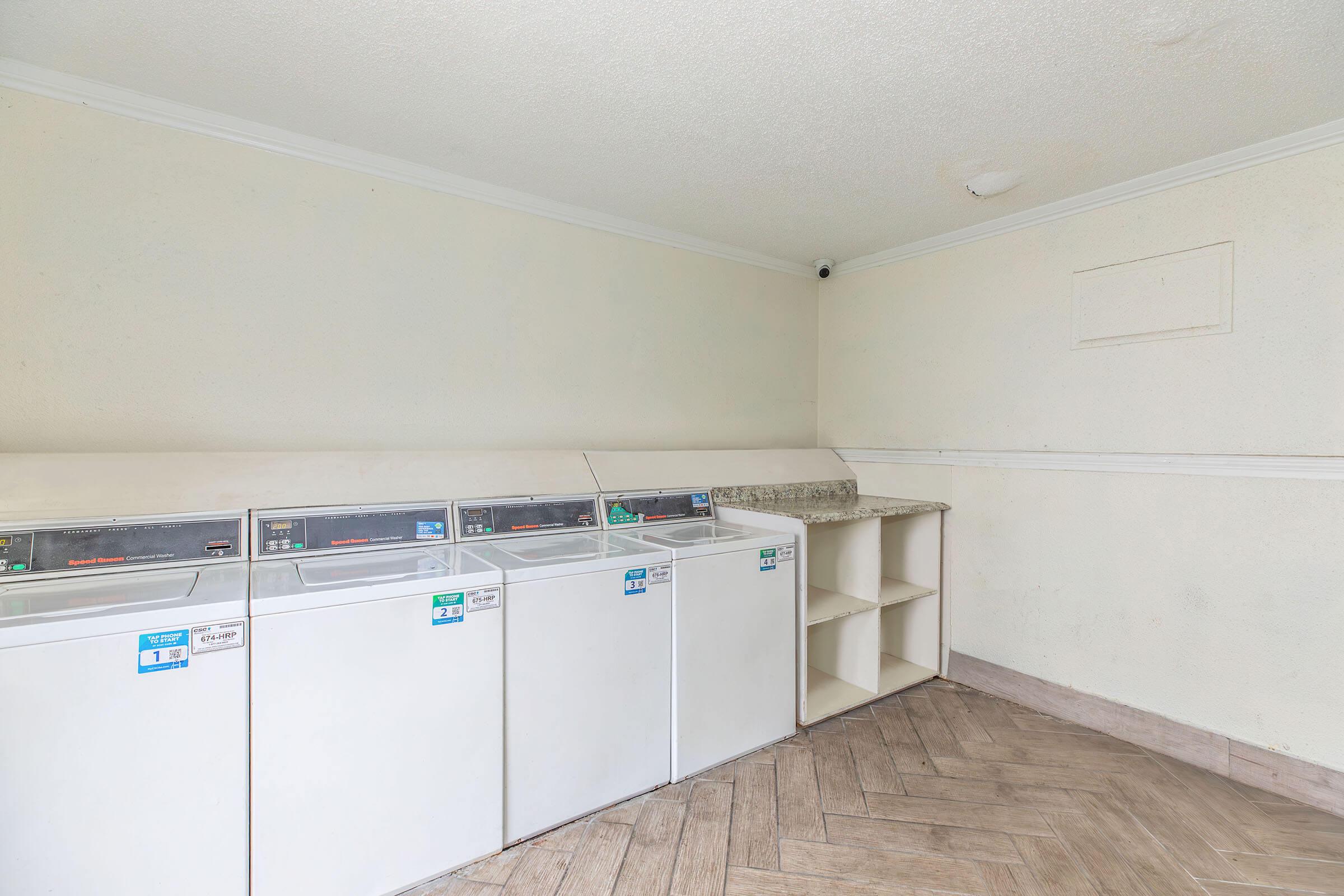 A clean, functional laundry room featuring three white washing machines lined up against a light-colored wall. There is a countertop alongside the machines and a shelf unit nearby. The flooring is a light wood pattern, and the room is well-lit.