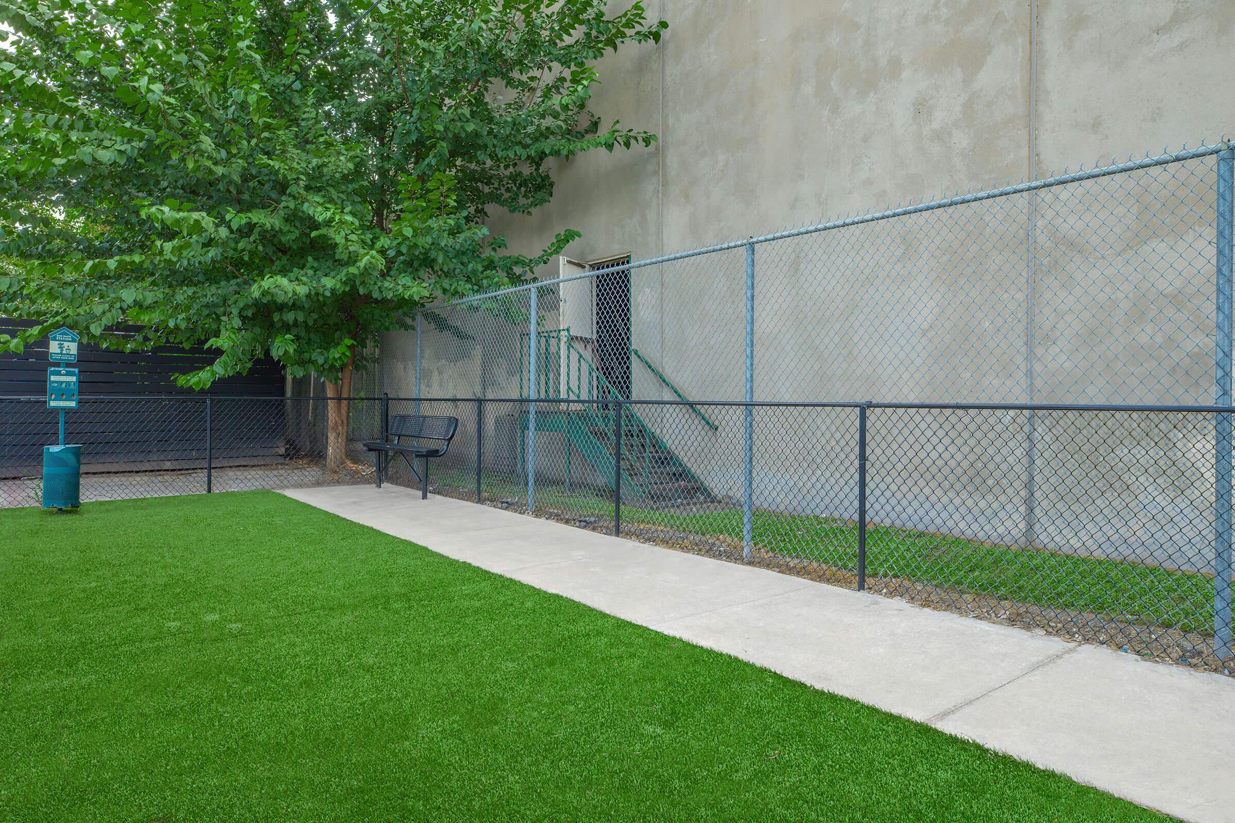 A grassy area with artificial turf, featuring a paved walkway along a chain-link fence. There’s a bench near the fence, and behind it, a green staircase leads to a door. A leafy tree provides shade, and a signpost stands nearby, marking the area. The wall in the background is gray and unadorned.