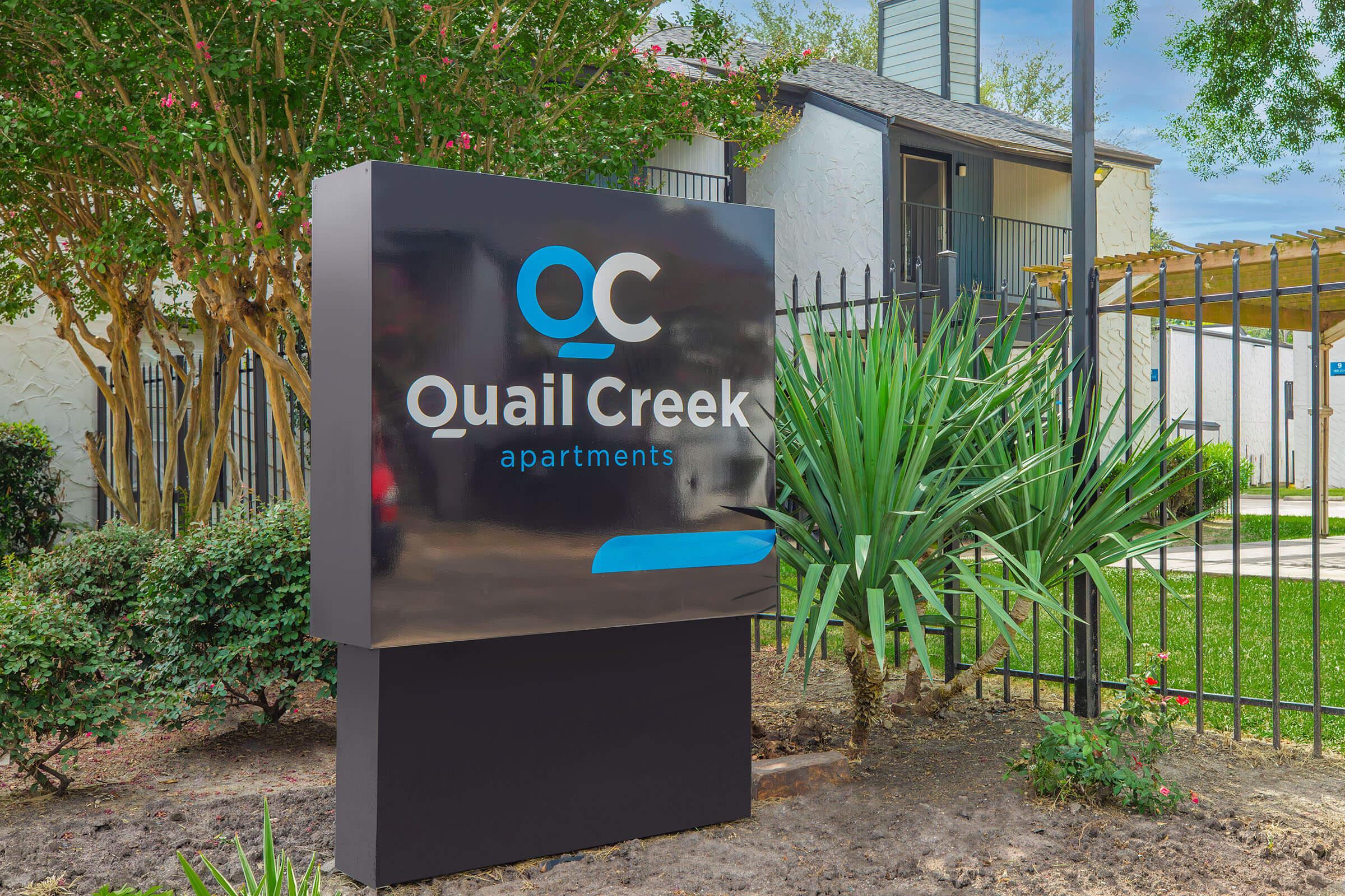 Sign for Quail Creek Apartments, featuring the logo "QC" above the name "Quail Creek apartments." The sign is modern and prominently displayed in front of a landscaped area with green plants and trees, along with an apartment building visible in the background.