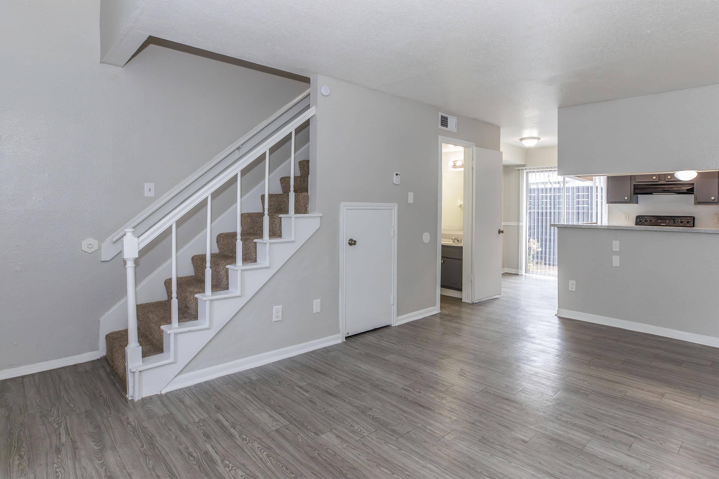 A modern living space featuring an open layout. A staircase leads to the upper level, while the main floor has a light wood-style floor. There is a small door under the stairs, and the area opens into a kitchen visible in the background, with natural light coming through a sliding door. Neutral walls create a spacious feel.