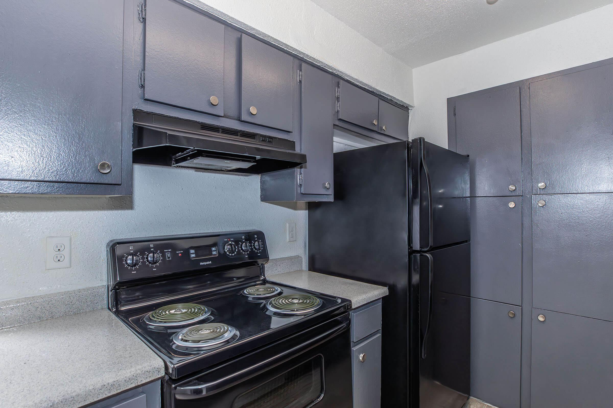 A modern kitchen with dark blue cabinets, a black refrigerator, and an oven with four burners. The countertop is light gray, and the kitchen has simple overhead cabinets, providing ample storage space. The design is sleek and functional, showcasing a clean and organized cooking area.