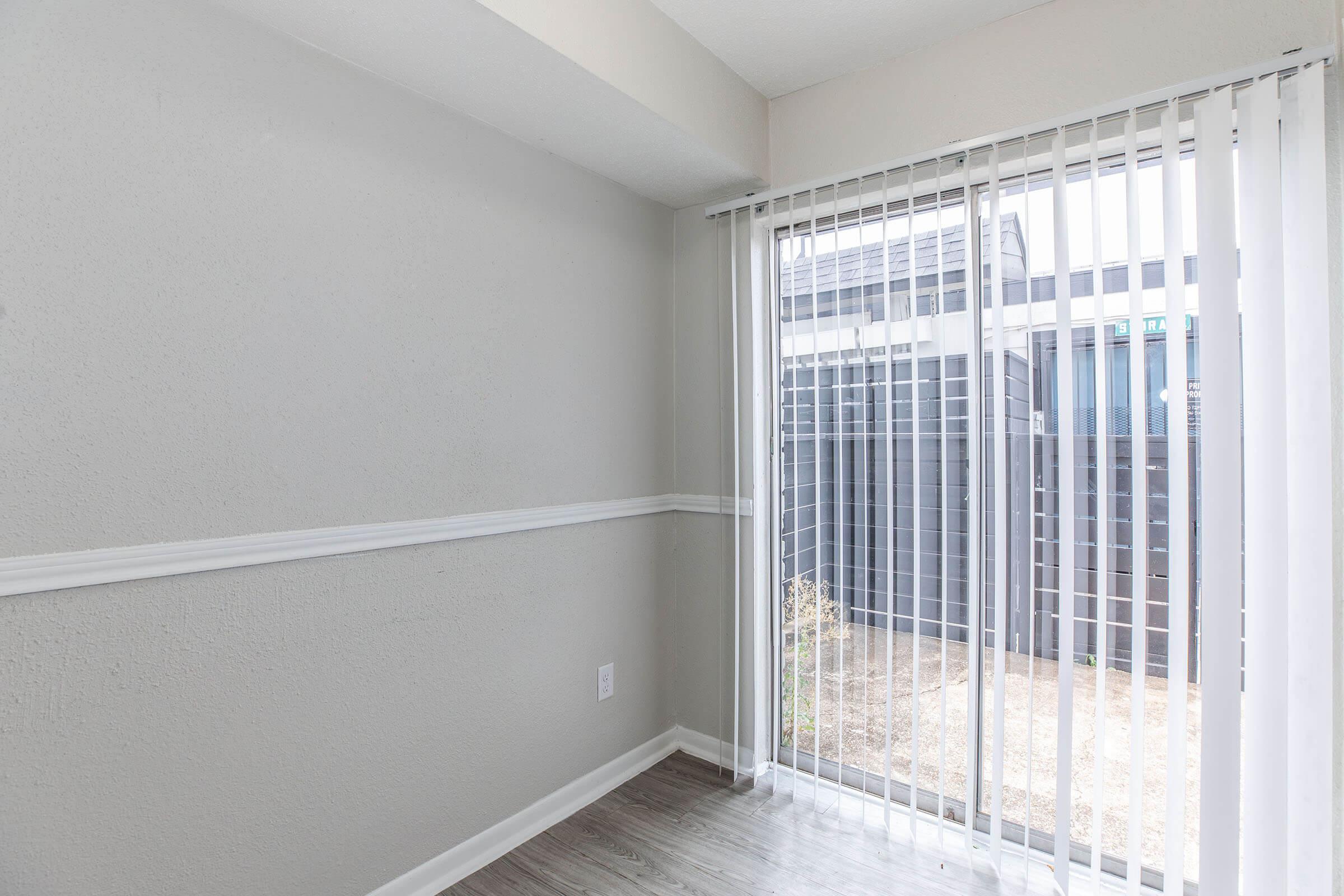 A clean, empty room featuring light gray walls and a window with vertical blinds leading to an outdoor area. The floor is light-colored wood, and there's a simple white baseboard. Natural light brightens the space, giving it a fresh and airy feel.