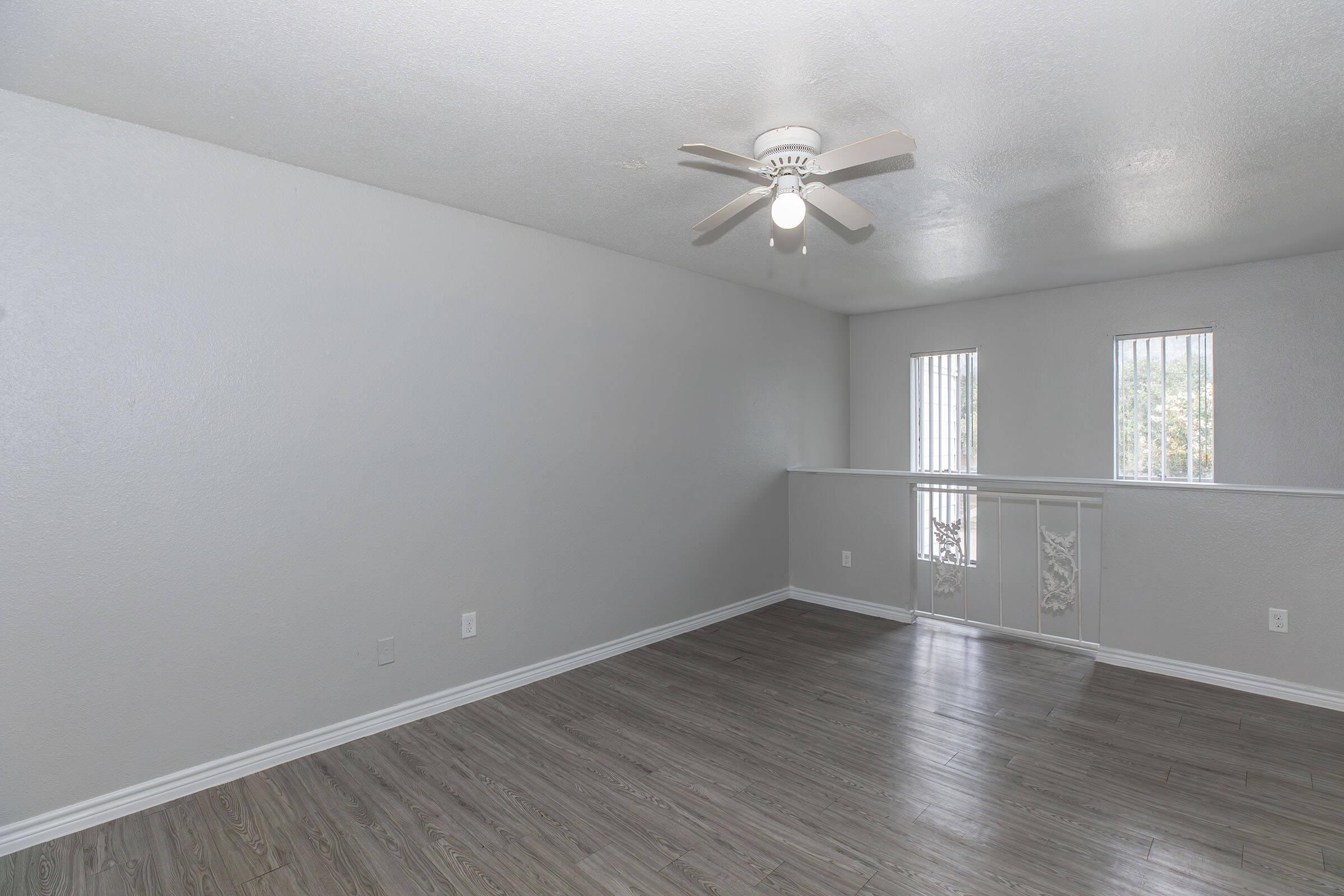 A spacious room with light gray walls and a ceiling fan. It features a wood-like floor and two small windows with blinds. A railing separates the room from a staircase area, creating an open and airy feel. The overall atmosphere is bright and inviting.