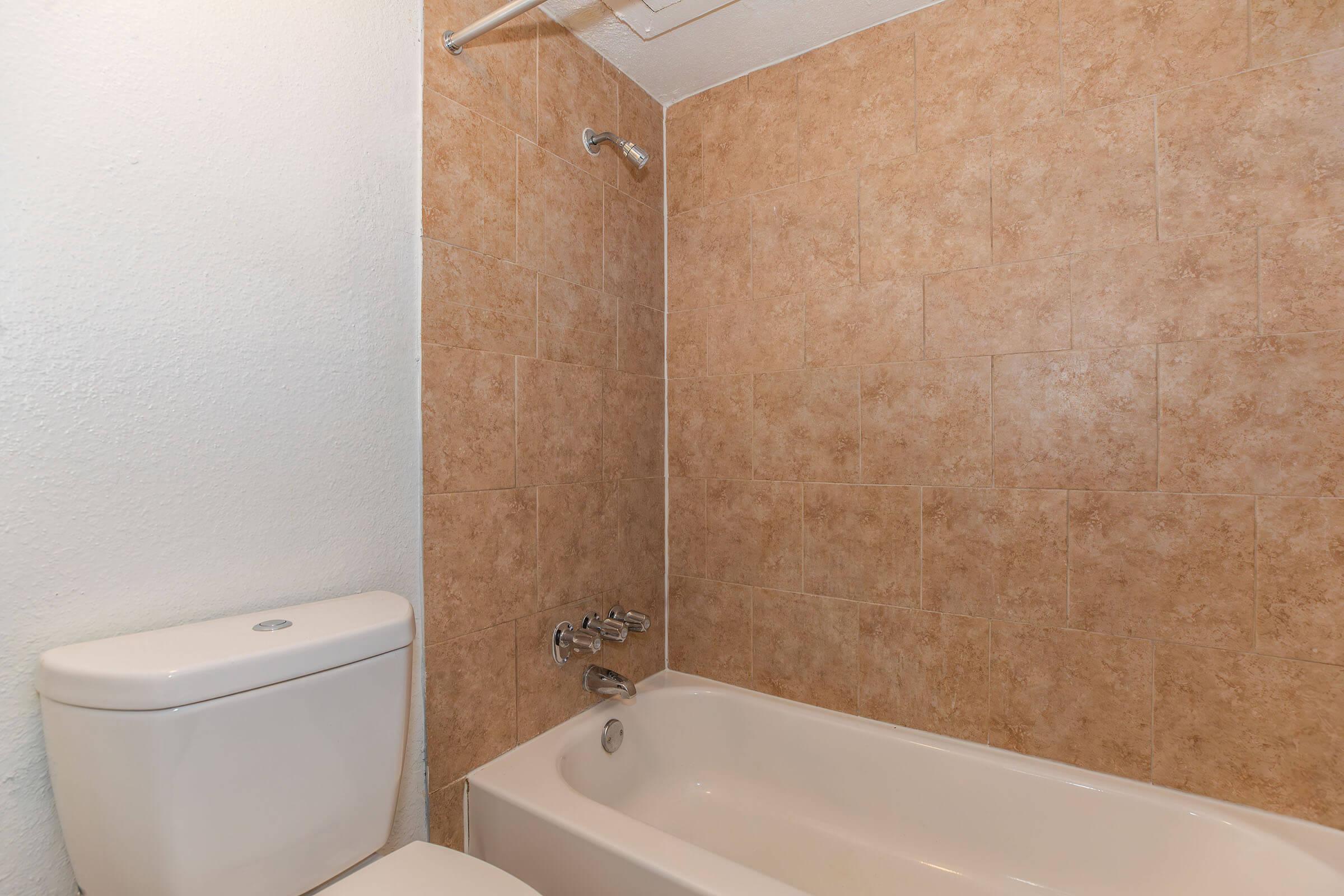 A clean bathroom featuring a beige tiled wall, a white bathtub, a chrome faucet, and a toilet. The layout is simple, with no additional decorations or items present, creating a minimalist and functional space.