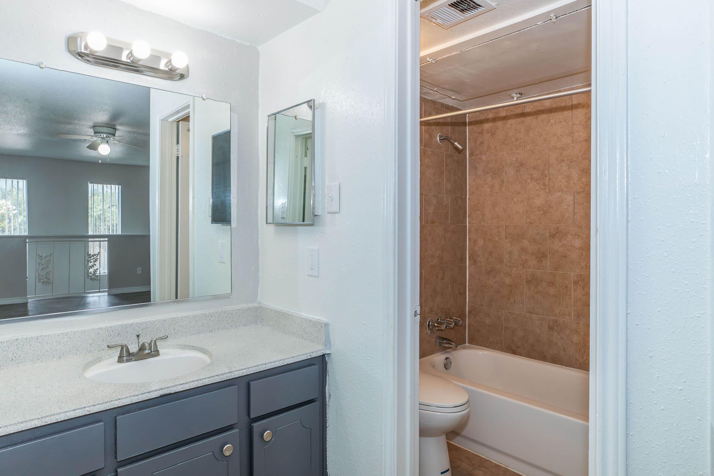 A well-lit bathroom featuring a modern vanity with a sink, a large mirror, and a small mirror on the wall. There is a bathtub with a showerhead, surrounded by tiled walls, and the decor includes light-colored walls and a ceiling fan visible through a doorway.