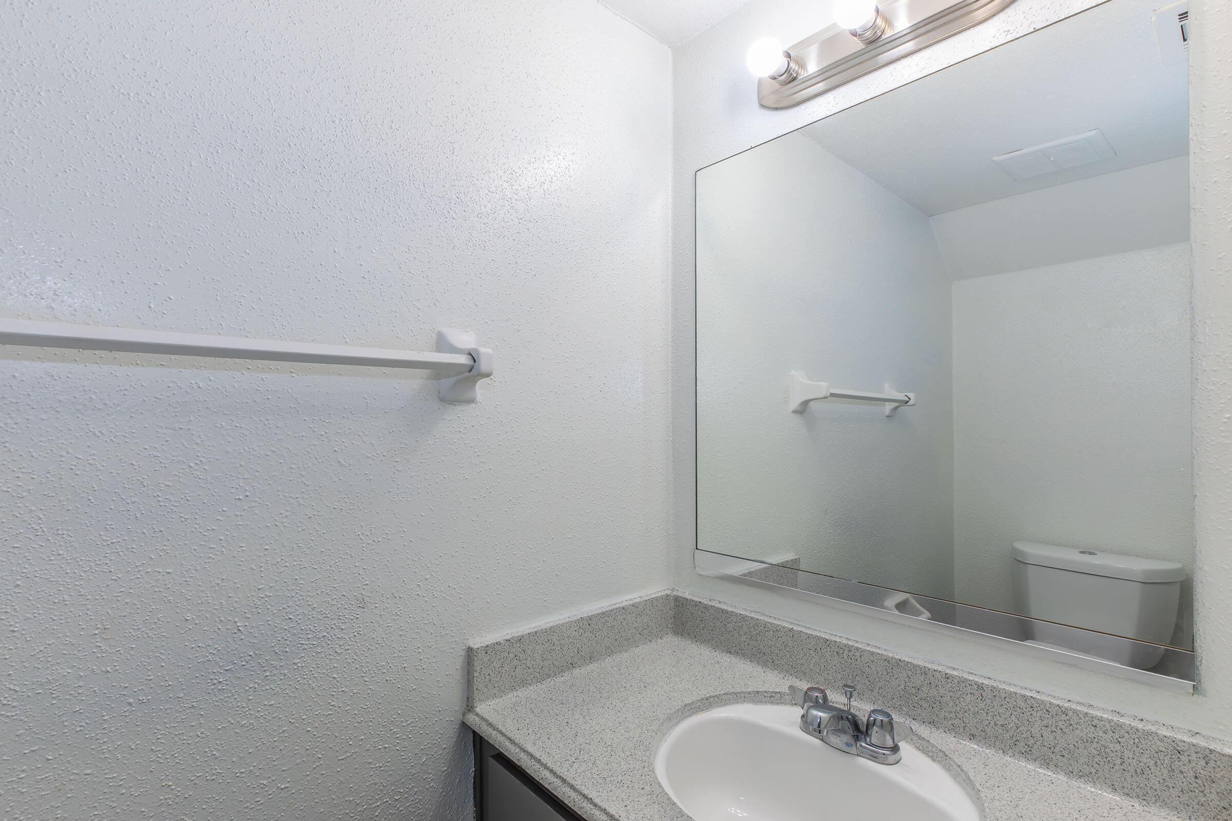 A clean, minimalist bathroom featuring a white wall, a small sink with a faucet, and a mirror above the sink. There is a towel bar mounted on the wall and a toilet visible in the reflection of the mirror. The overall color scheme is light and neutral, contributing to a simple and functional design.