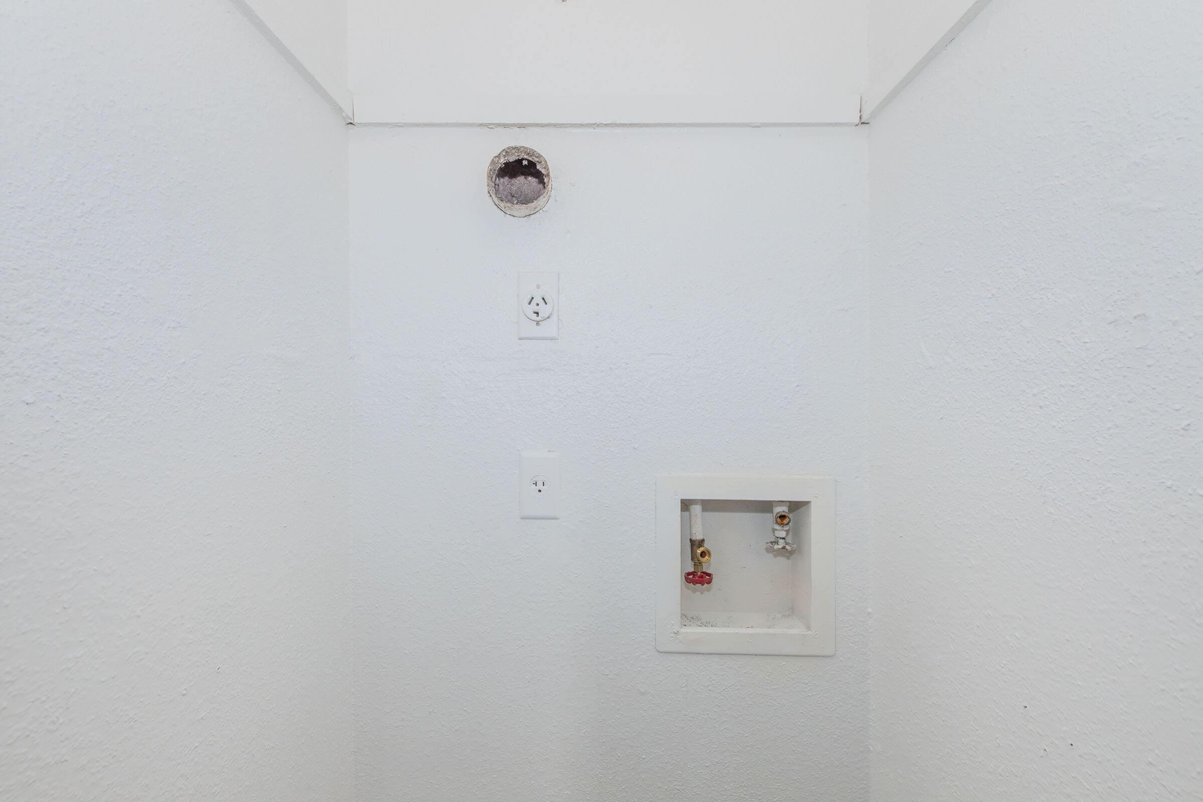 A narrow, white-walled space with a small circular vent on the left and two electrical outlets above. Below, there's a recessed area containing two valves, one red and one silver, mounted in a white frame. The overall appearance is clean and minimalistic, with no other distinguishing features.