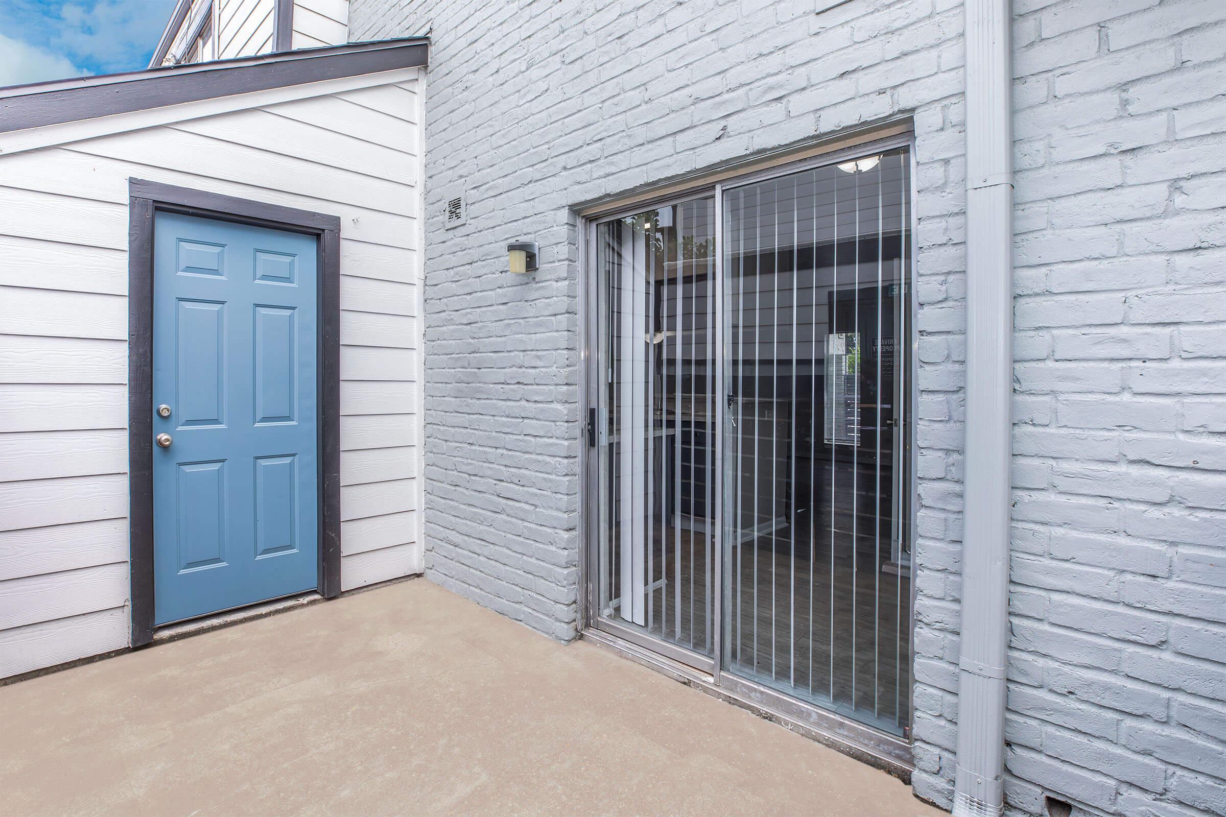 Exterior view of a home featuring a blue door on the left leading to a room and a sliding glass door on the right, with a brick wall and concrete floor in the area between them. Natural light filters through the sliding door, highlighting the interior space beyond.