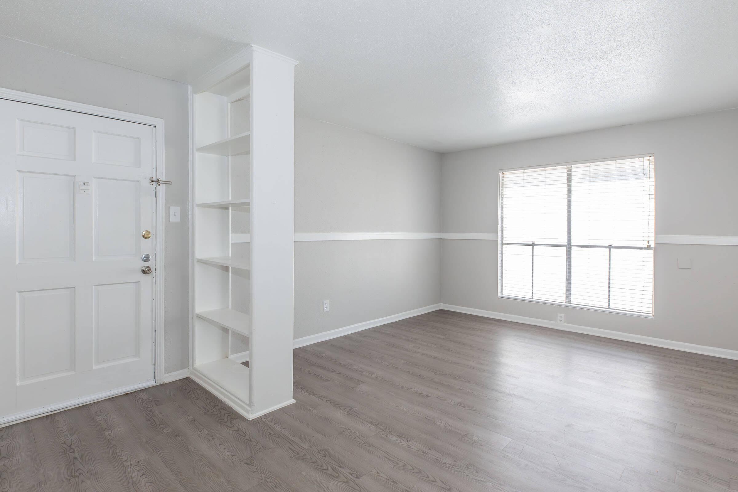 A bright, empty living room featuring light gray walls and laminate flooring. The room includes a door on the left, a built-in bookshelf on the right, and a large window with blinds that allows natural light to fill the space. The overall feel is modern and minimalistic.