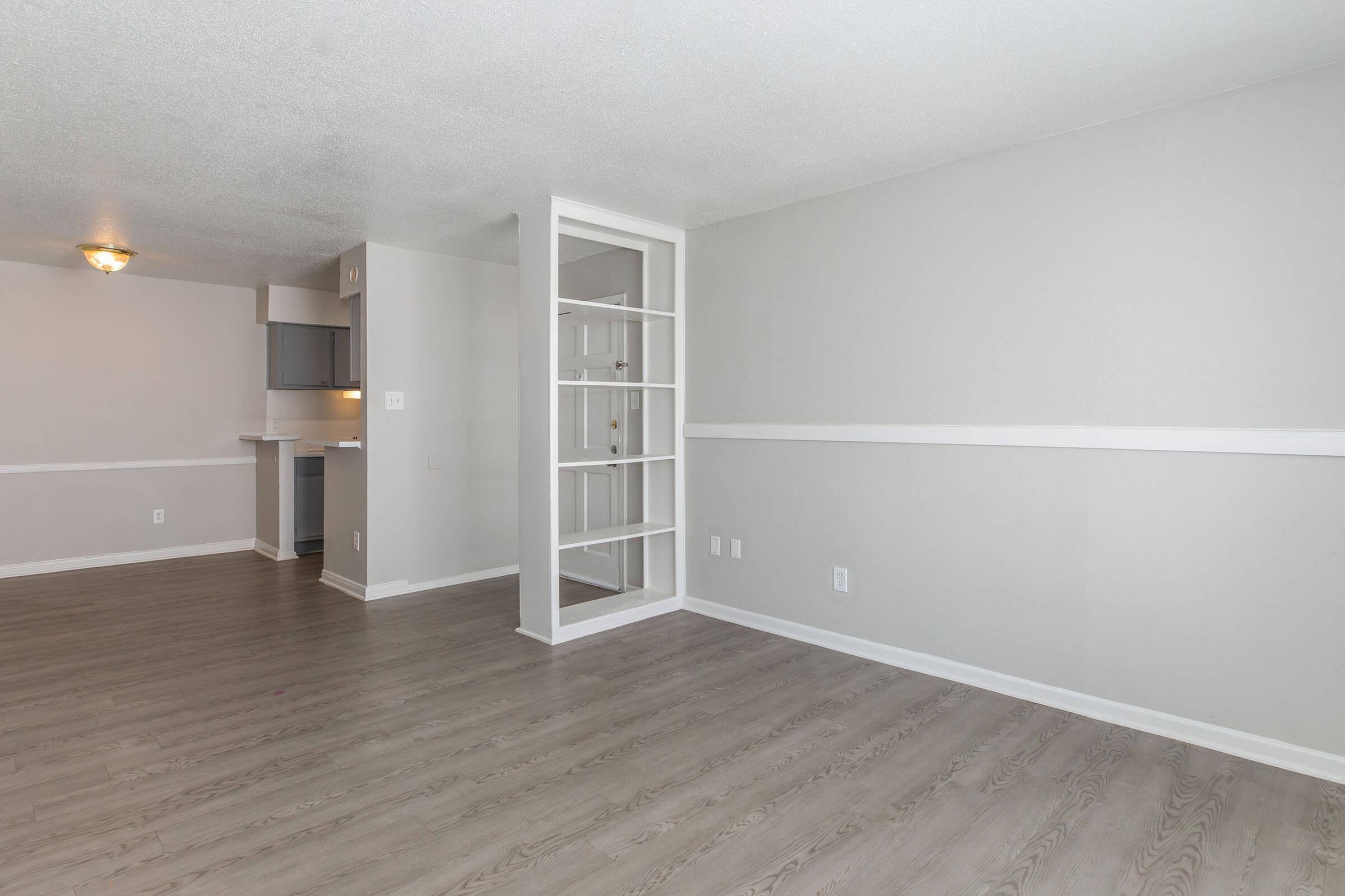 A light, airy interior of an apartment living space featuring smooth gray walls, new laminate flooring, and an open layout. A partition with glass panels separates the living area from the kitchen, which is partially visible in the background. Natural light from a ceiling fixture adds warmth to the space.