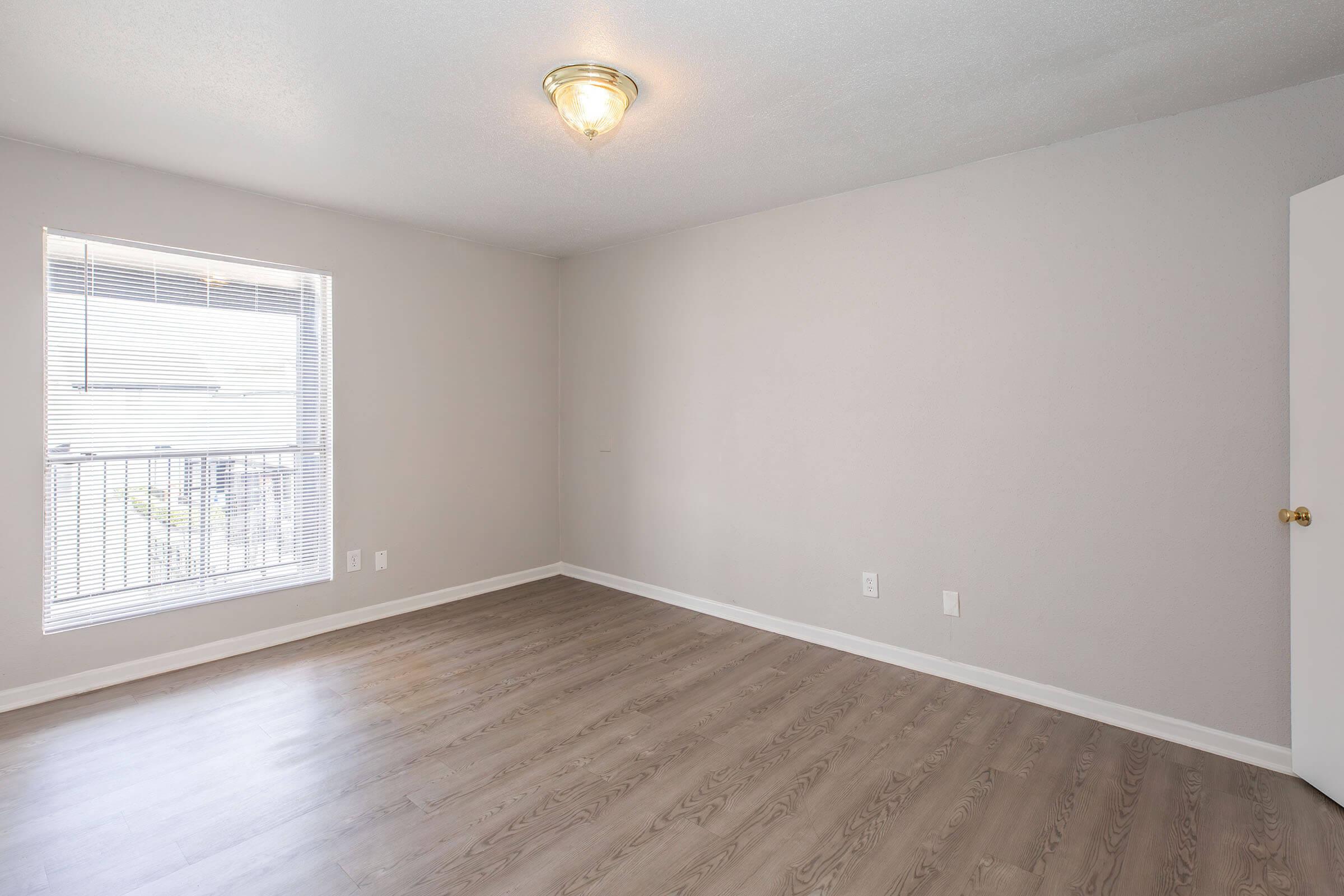 A spacious, empty room featuring light gray walls, a wooden floor, and a ceiling light fixture. There is a window with horizontal blinds allowing natural light. A door is visible on the right, leading to an adjacent area. The overall atmosphere is clean and well-lit, suitable for various purposes.