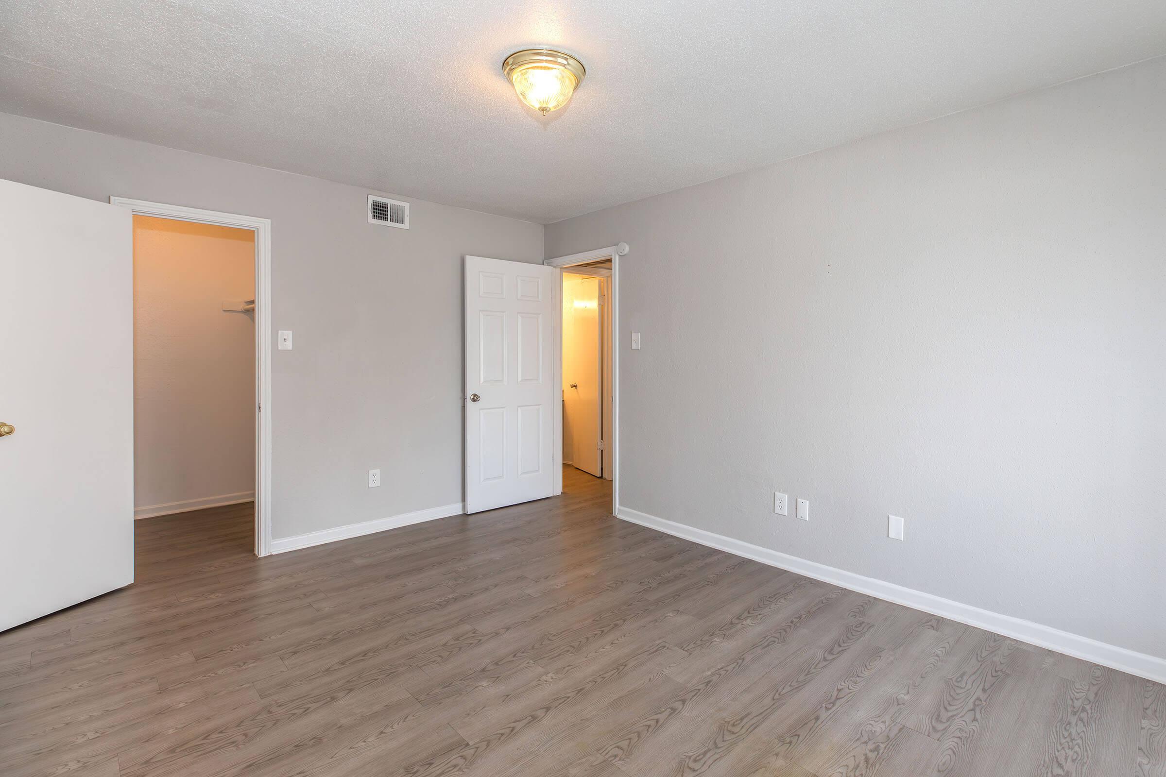 A simple, empty room with light gray walls and a hardwood floor. There is a ceiling light fixture, a closed white door on the left leading to a closet, and an open door on the right leading to another space. The room is bright and uncluttered, suggesting potential for decoration or furnishing.
