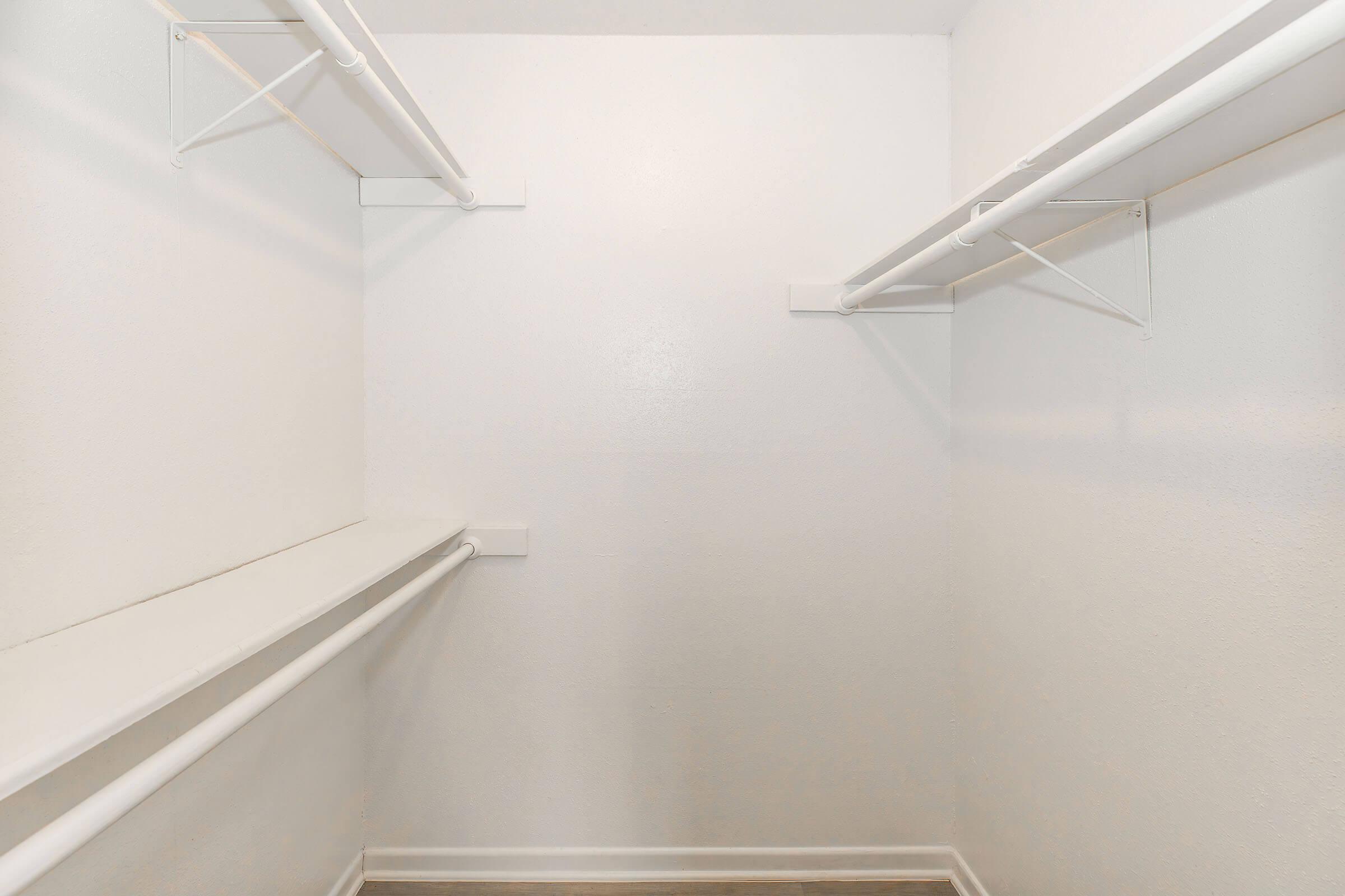 Empty closet interior featuring white walls and two white hanging rods. The floor is a light wood finish, and there is a shelf along one side. The space appears clean and organized, with no visible items or clutter.