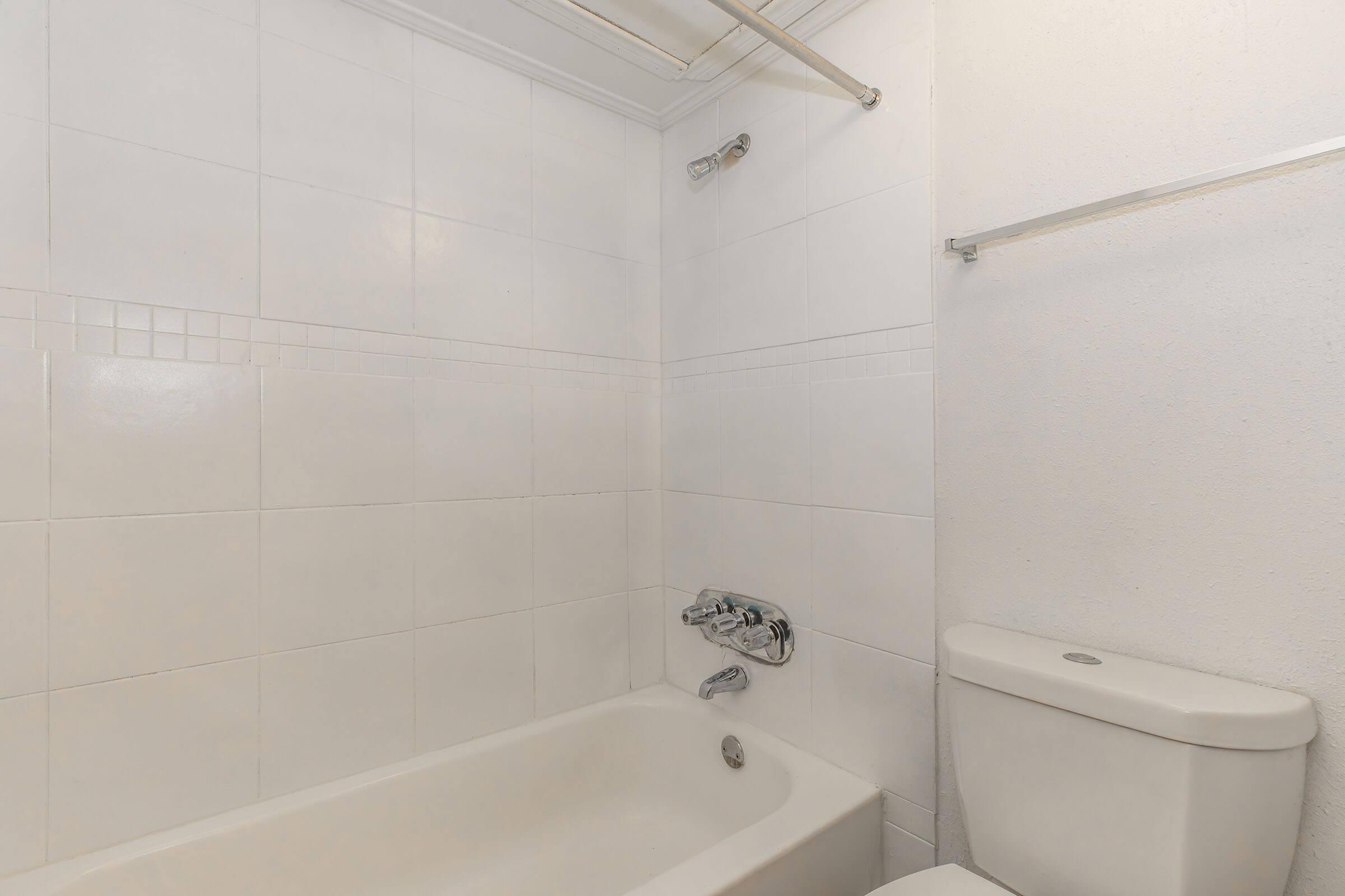 Interior view of a bathroom featuring a clean, tiled wall with a bathtub on the left, a chrome faucet, and a showerhead. On the right, there is a white toilet. The overall design is minimalistic and bright, with neutral colors dominating the space.