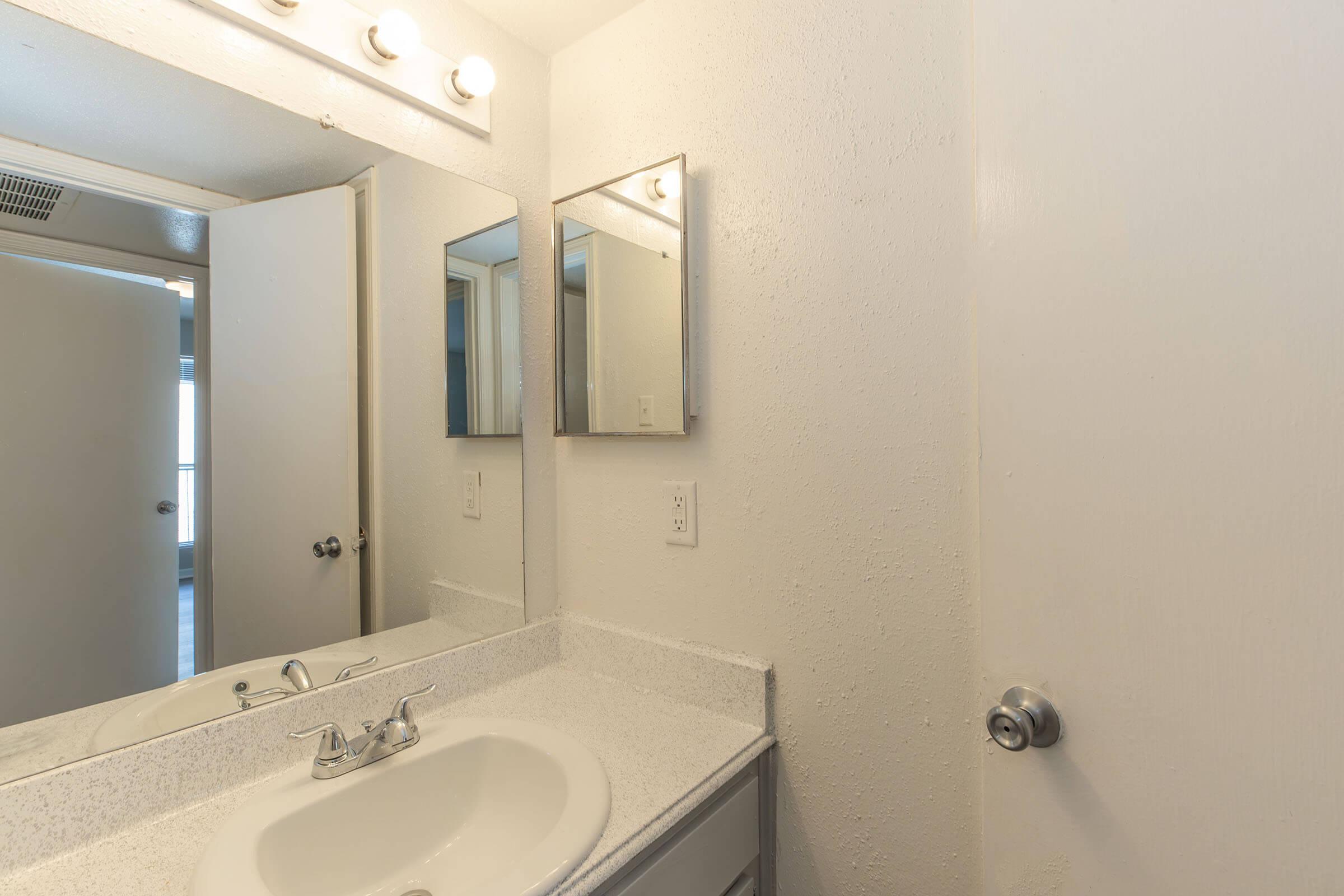 A small bathroom featuring a white countertop with a sink, a large mirror above the sink, and bright overhead lights. The walls are light-colored, and there is a partially open door leading to another room. A bathroom cabinet is visible beside the sink.