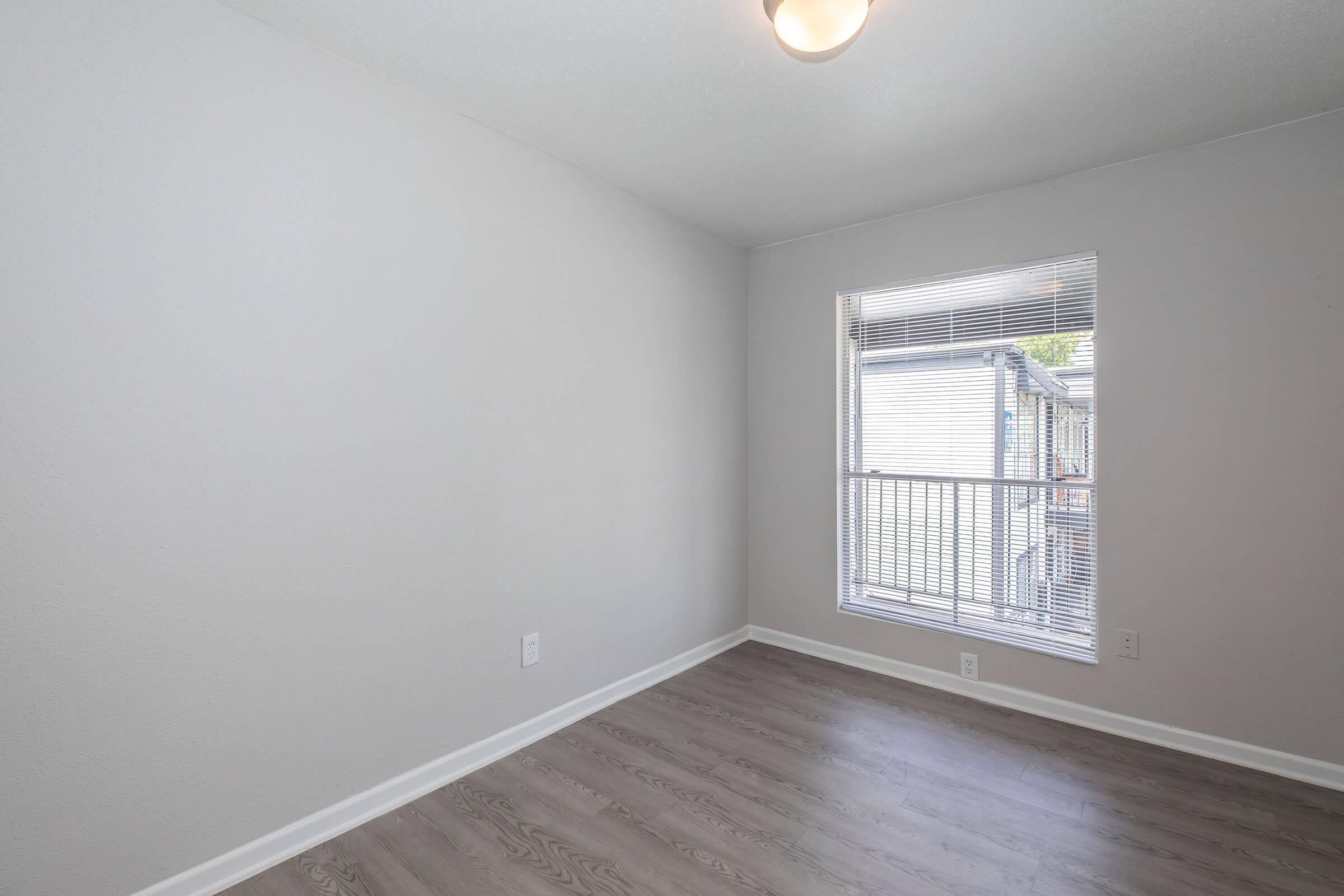 Empty room with light gray walls, a window covered by blinds, and wooden flooring. The space is well-lit with a ceiling light fixture, showcasing a clean and minimalist design. The room has no furniture or decorations, emphasizing its openness and potential for personalization.