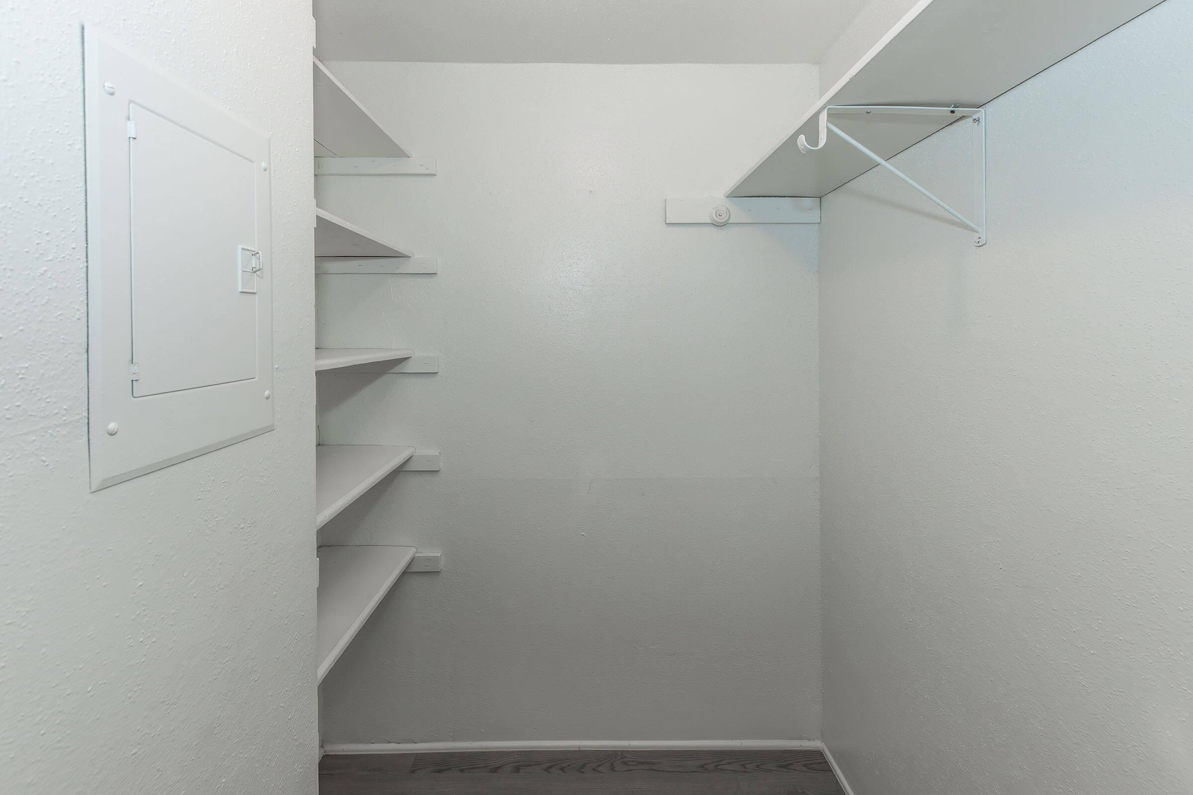 A clean, empty closet featuring white walls and shelves. There are built-in shelves on the left side and a hanging rod on the right side. The floor is a light wood finish, creating a bright and spacious appearance suitable for storage.