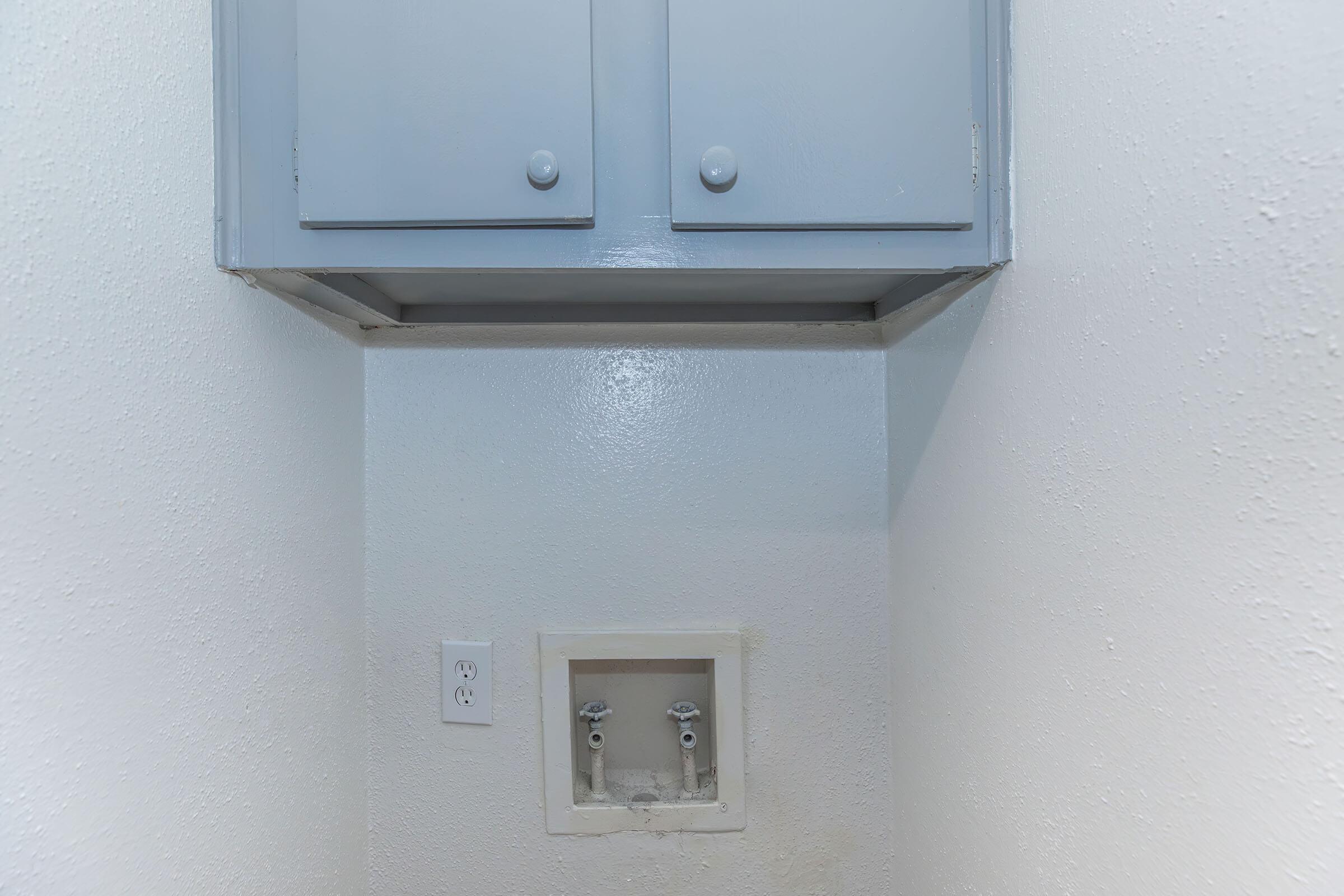 A small, empty laundry space featuring a light gray cabinet mounted above a white wall. Below the cabinet, there is a rectangular opening with water hookups, and an electrical outlet on the side wall. The walls are painted white with a textured finish.