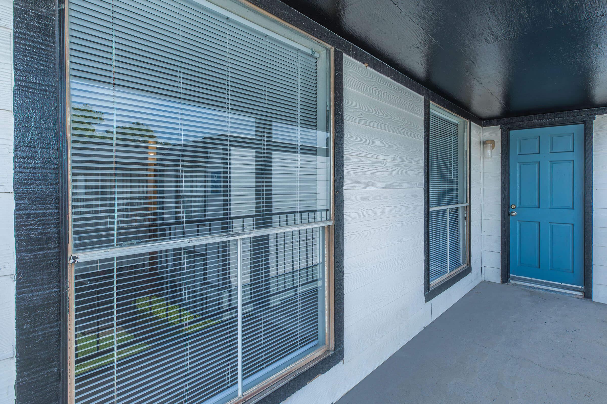 A view of a residential porch featuring two large windows with blinds and a bright blue front door. The walls are white with black trim, and the ceiling above is painted black. The porch floor is plain and concrete, providing an inviting entrance to the home.