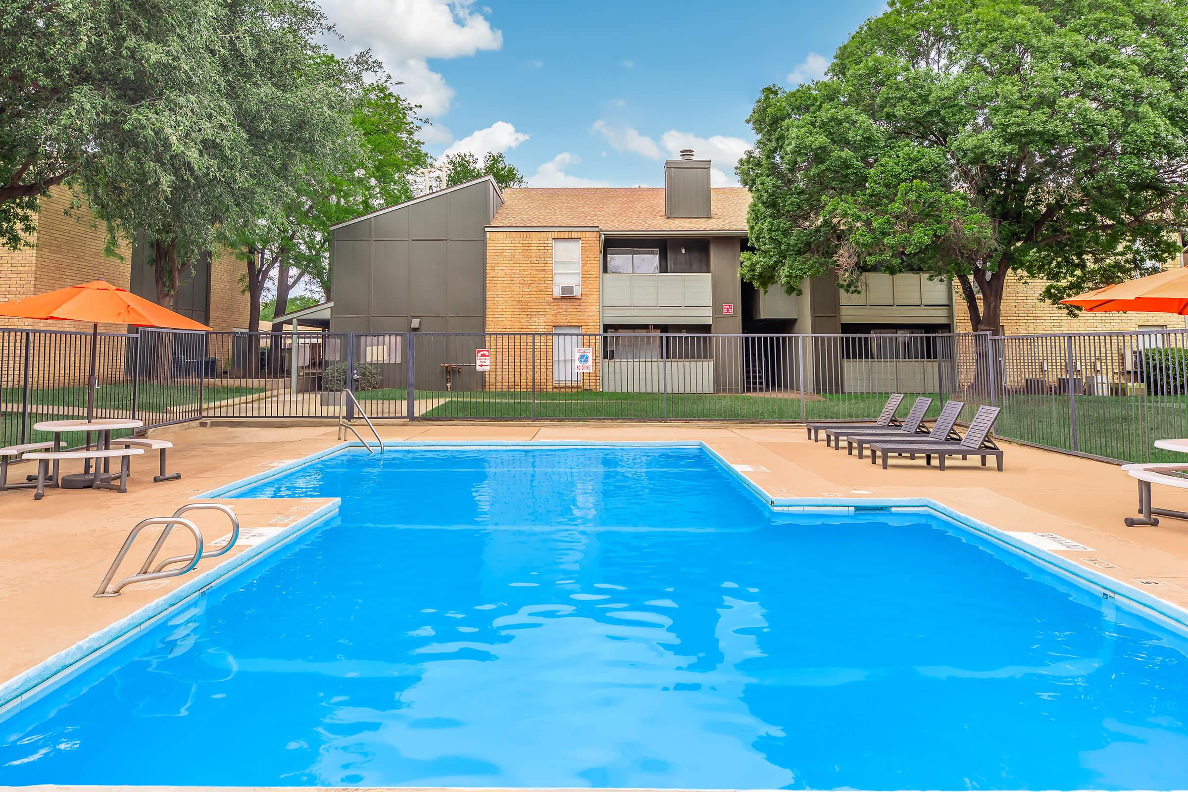 A bright outdoor swimming pool surrounded by lounge chairs and orange umbrellas, with green trees and an apartment building in the background. The clear blue water reflects the sky and provides a relaxing atmosphere.