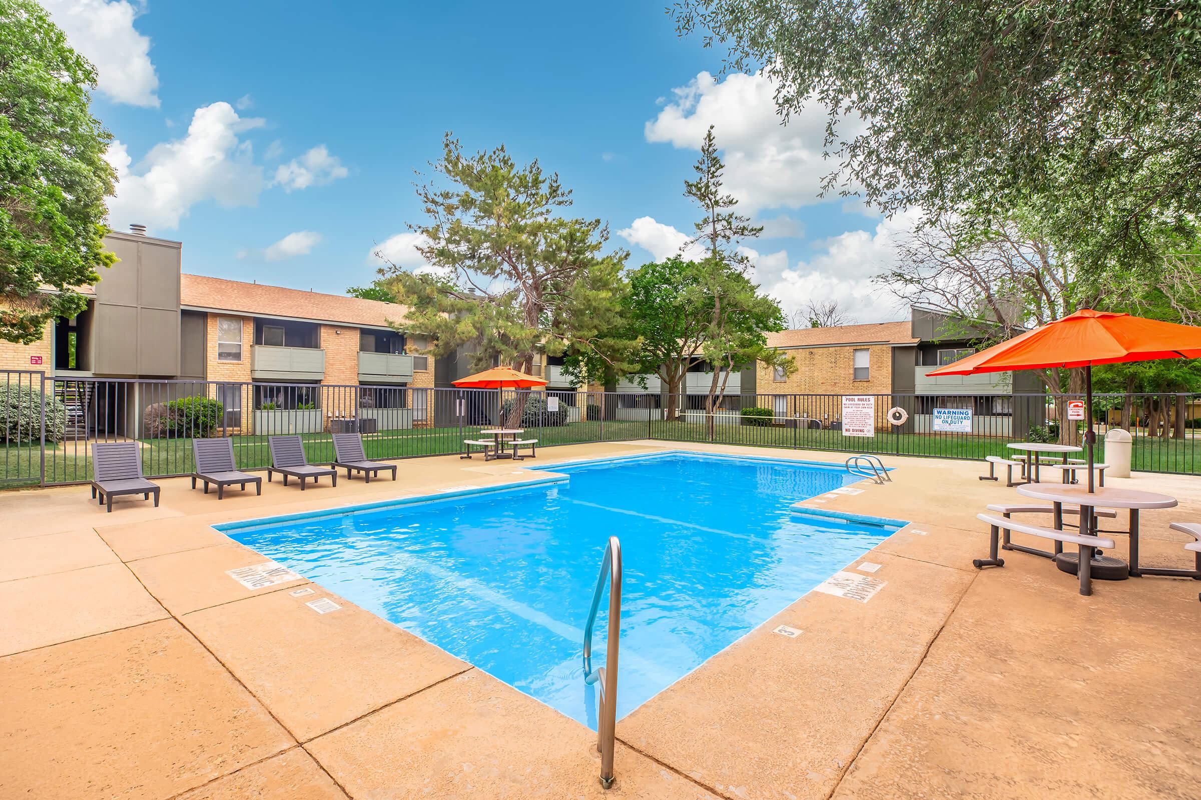A clear blue swimming pool surrounded by lounge chairs with orange umbrellas. In the background, there are brick buildings and trees under a bright blue sky with fluffy white clouds. The pool area appears inviting and well-maintained, ideal for relaxation and socializing.