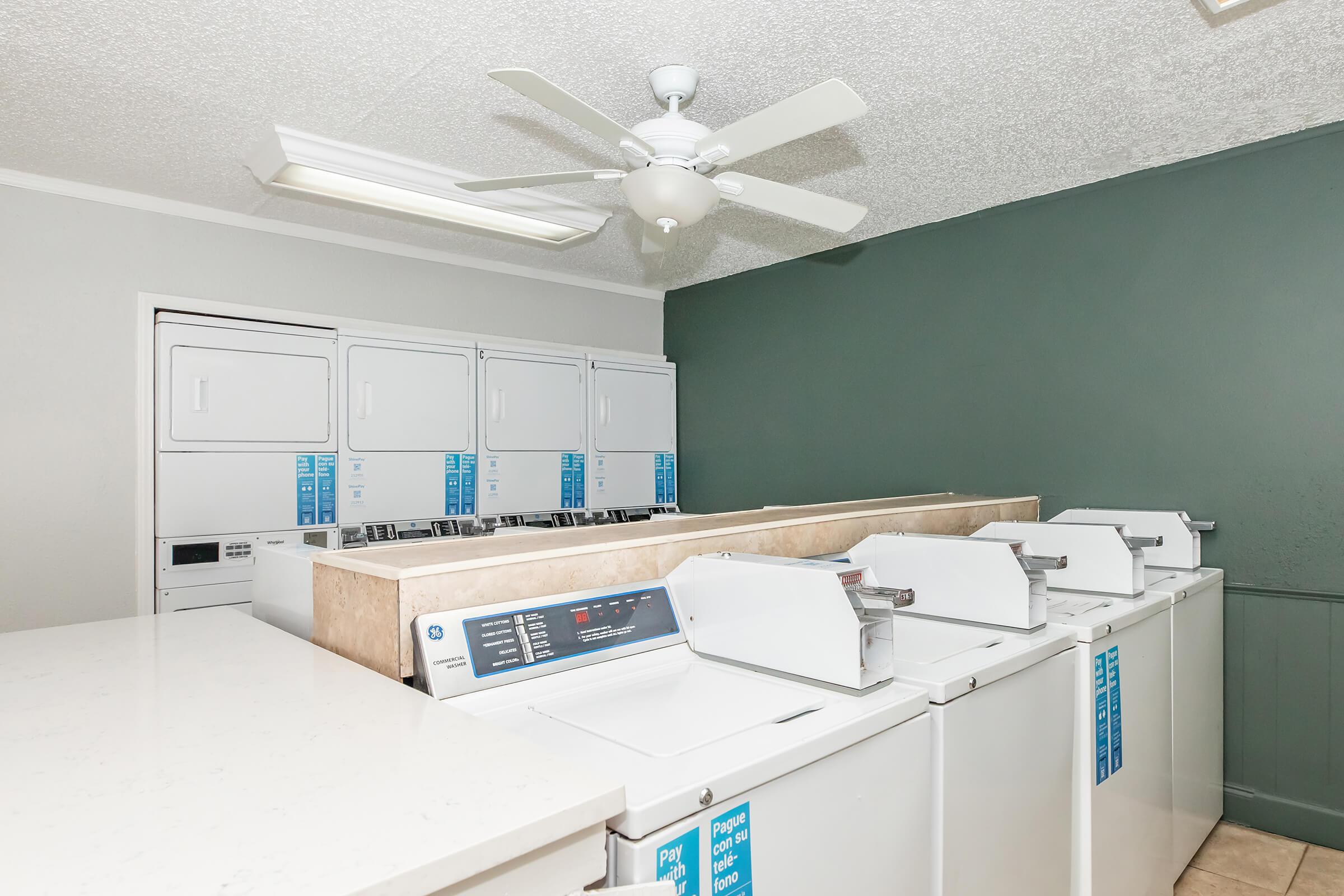 A clean and well-lit laundry room featuring several washing machines and dryers. There are multiple stacked laundry units against the wall, and a ceiling fan is visible. The walls are painted a muted green, and there is a countertop in the foreground.