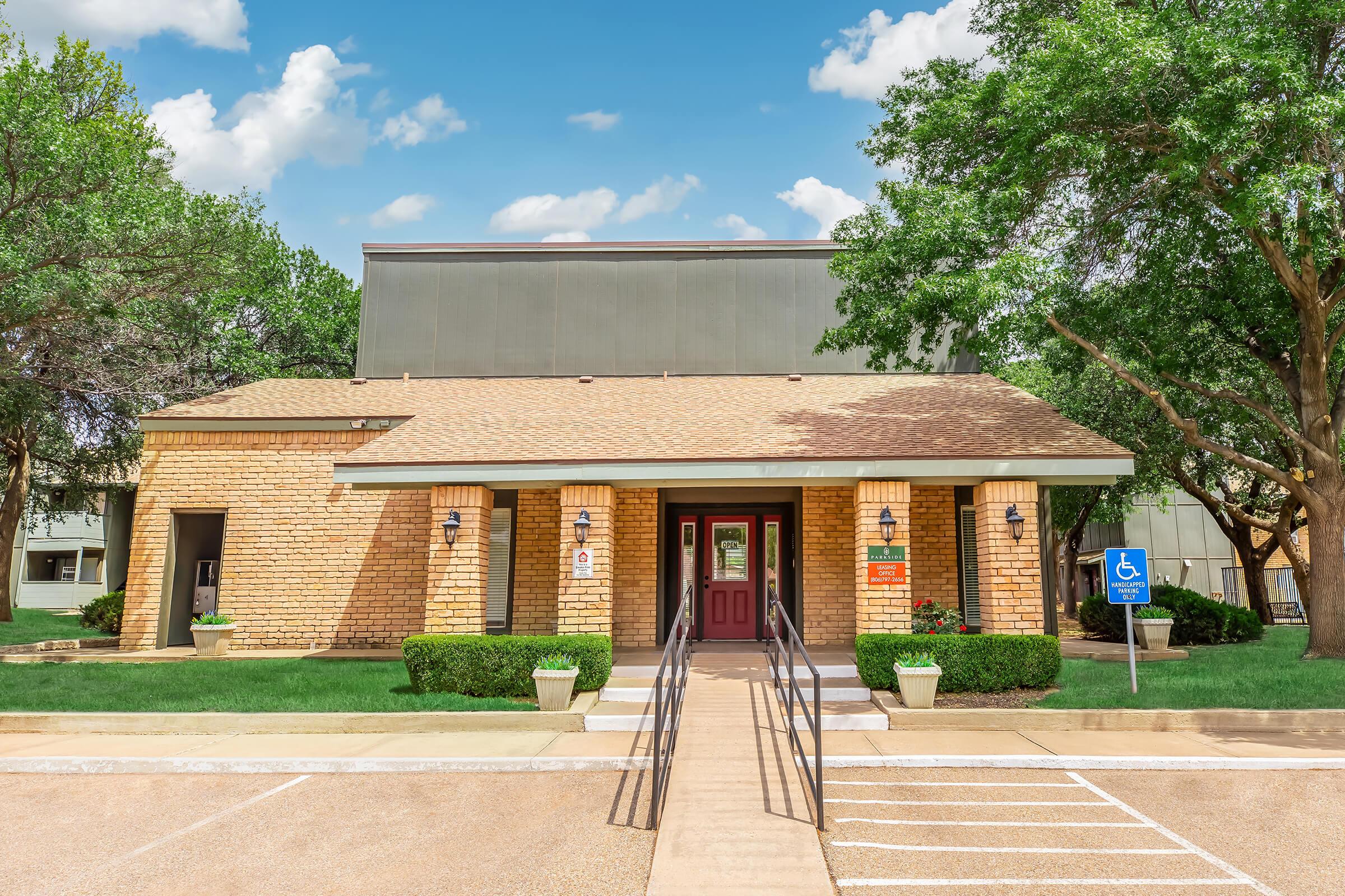 Single-story stone building with a sloped roof, featuring a red door and large windows. The entrance is accessible via a paved walkway with handrails. Surrounding greenery includes shrubs and trees under a bright blue sky with scattered clouds. A parking lot is visible in front, including spaces for handicapped access.