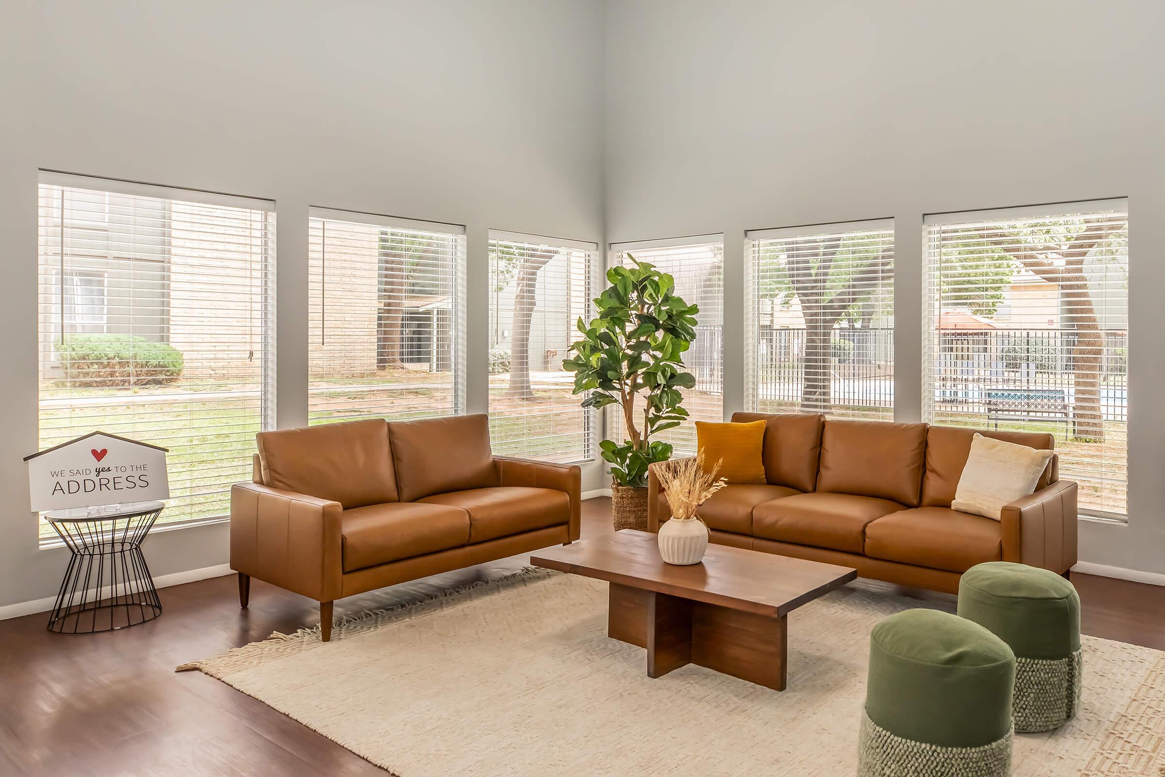 A bright living room featuring two brown leather sofas facing each other, a wooden coffee table in the center, and a green plant in a pot. Large windows provide ample natural light and a view of an outdoor area. A decorative sign and green upholstered stools add a cozy touch to the space.