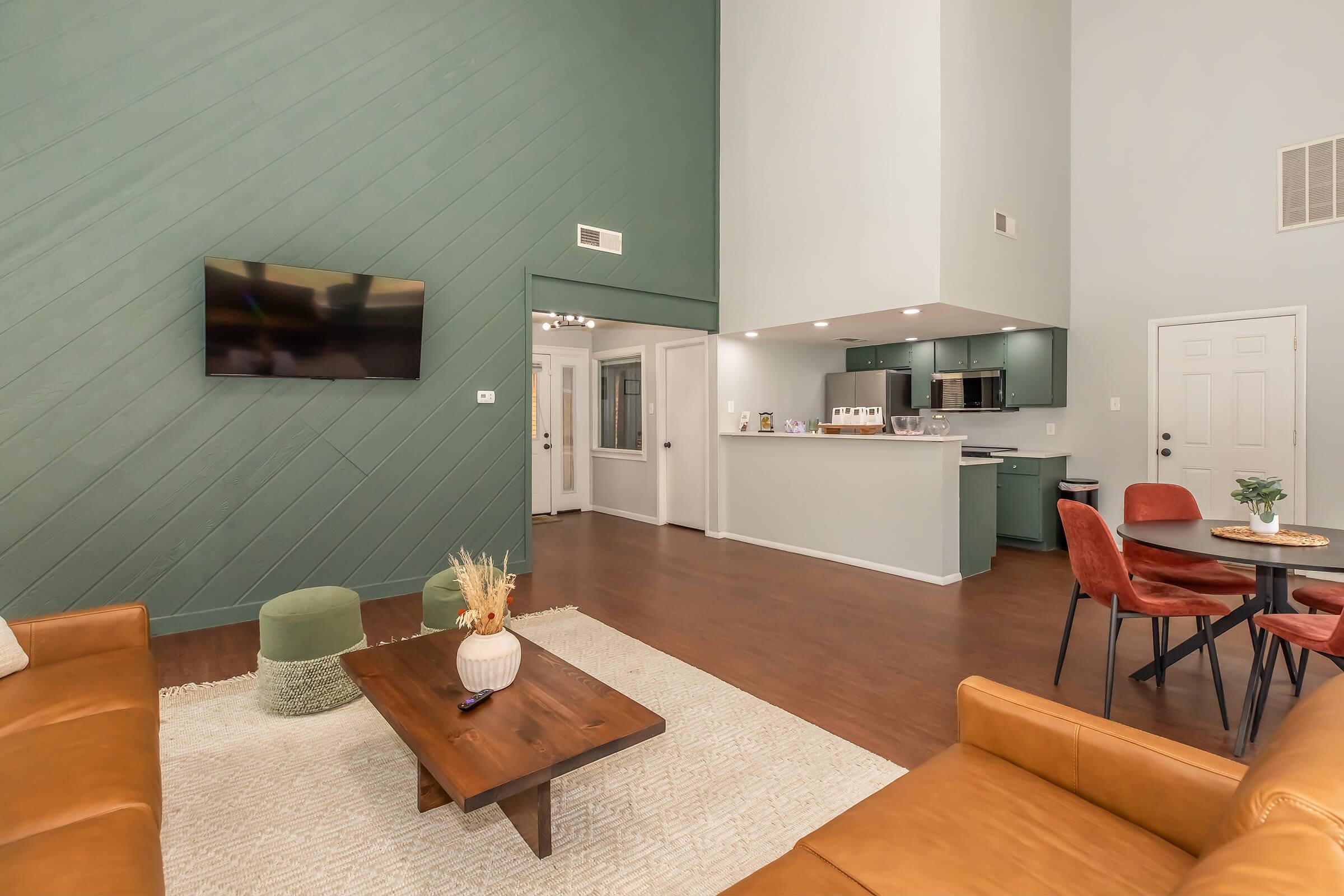 A spacious and modern living area featuring a large wall-mounted TV above a stylish brown leather sofa. The room has a green accent wall, a round dining table with four chairs, and a kitchen with an open layout. Natural light illuminates the space, creating a warm and inviting atmosphere.