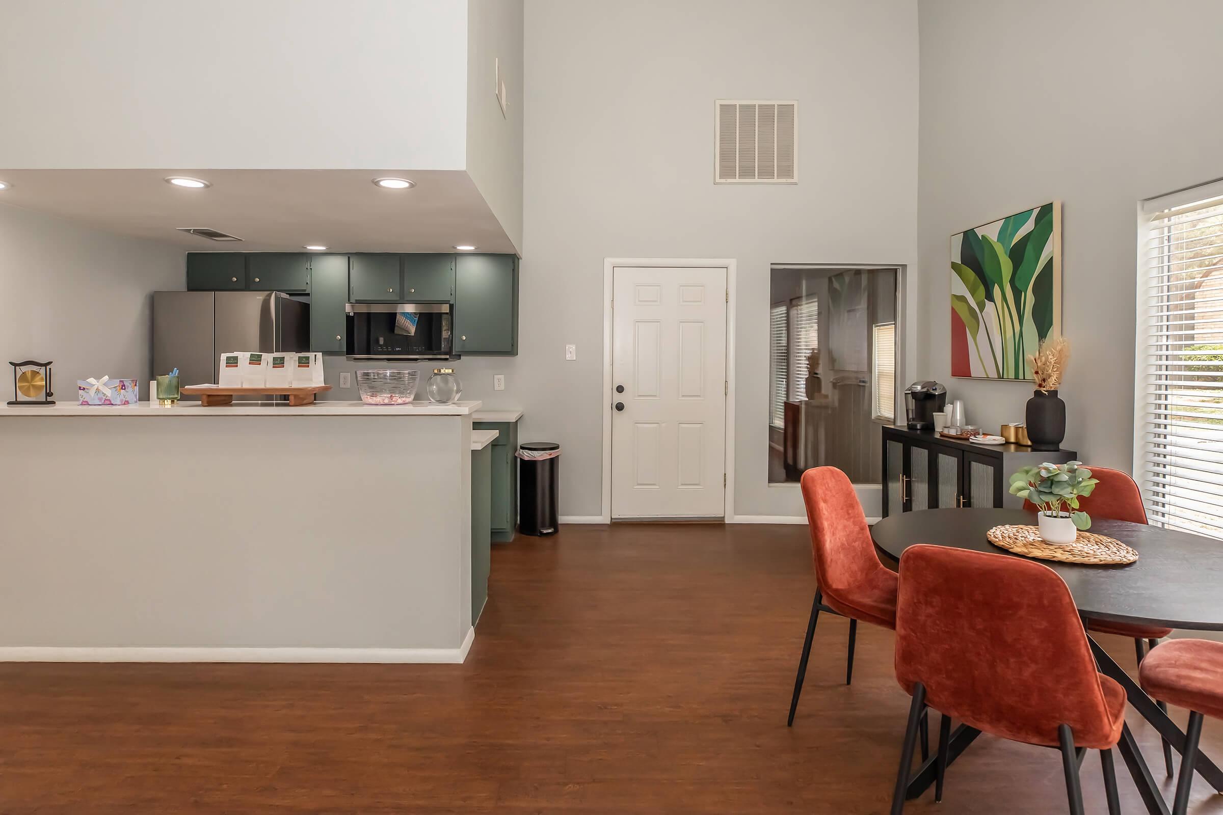 Modern kitchen and dining area featuring a sleek gray and green color scheme. The kitchen has stainless steel appliances and a breakfast bar. There’s a dining table with orange chairs, a door leading outside, and a well-lit ambiance created by natural light through the windows. Decorative plants add a cozy touch.