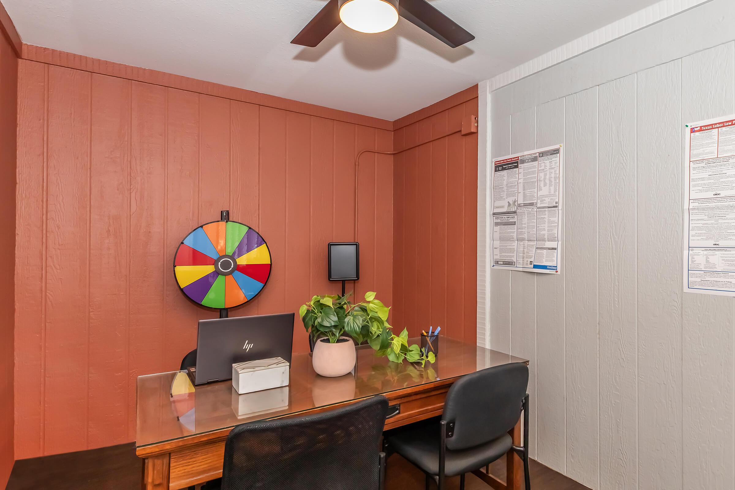 A cozy office space featuring a wooden desk with a laptop, an indoor plant, and a colorful spinning wheel on the wall. The walls are painted in contrasting colors, with one side in terracotta and the other in light gray. A small screen is mounted on the wall, and there are posters or documents displayed nearby.