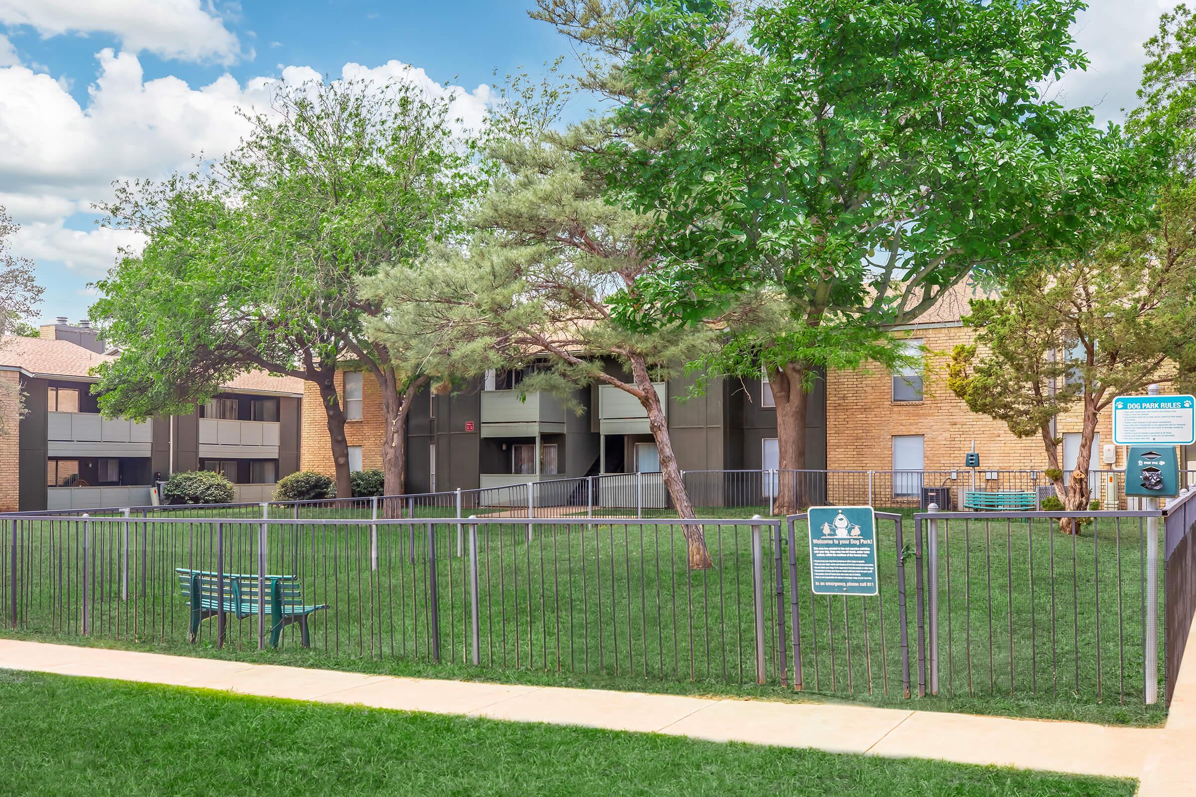 A grassy area enclosed by a black fence, featuring a green bench. Surrounding the space are several trees and a two-story apartment building in the background. The sky is partly cloudy, creating a bright and inviting atmosphere. A sign is visible near the fence, likely providing information about the area.