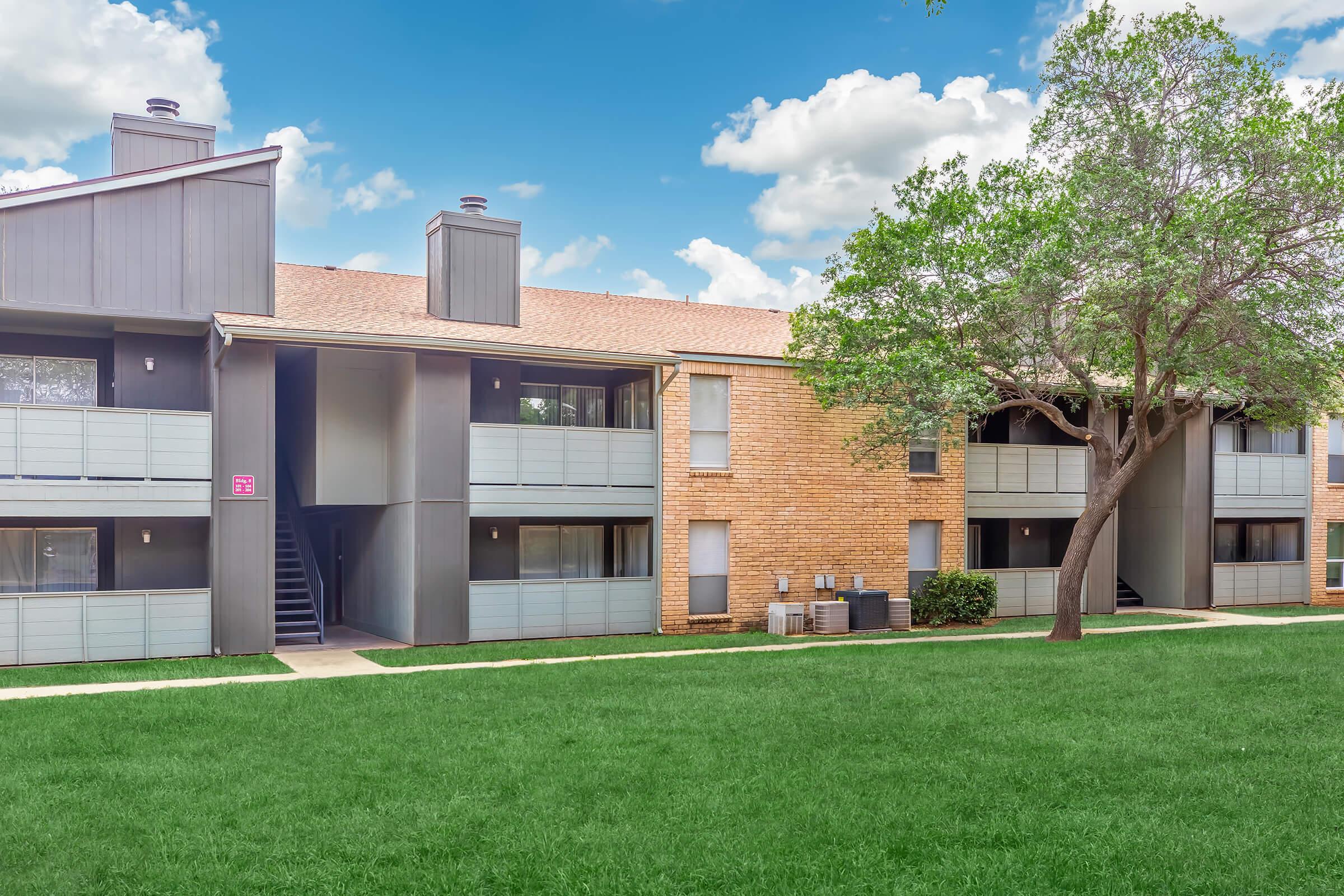 A two-story apartment building featuring a mix of brick and gray siding. The building has balconies and staircases leading to the upper level. A well-maintained green lawn with trees decorates the front, under a bright blue sky with scattered clouds.