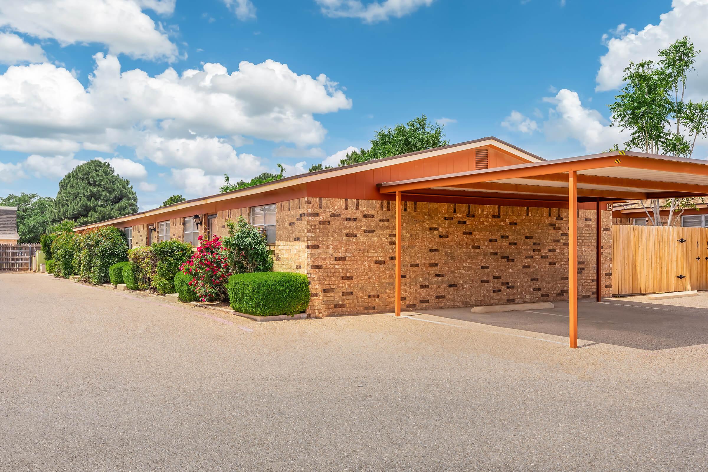 A brick building with a flat-roof carport, surrounded by well-maintained shrubs and flowers. The sky is bright with fluffy white clouds, and there is a gravel driveway leading to the structure. Trees and a wooden fence are visible in the background, creating a tranquil residential setting.