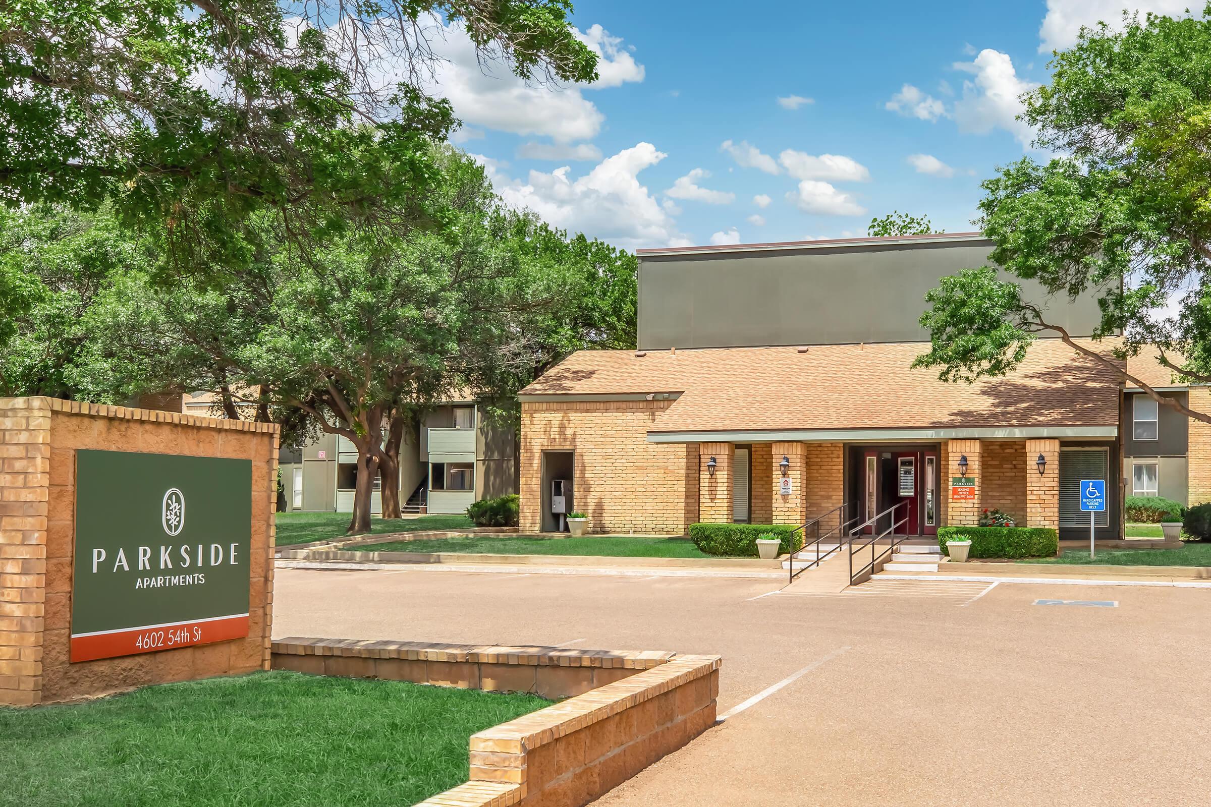 A brick building with a green sign reading "Parkside Apartments" at the entrance. The two-story apartment complex is surrounded by trees and grassy areas, with a clear blue sky above. There are accessible parking spaces and a welcoming walkway leading to the entrance.