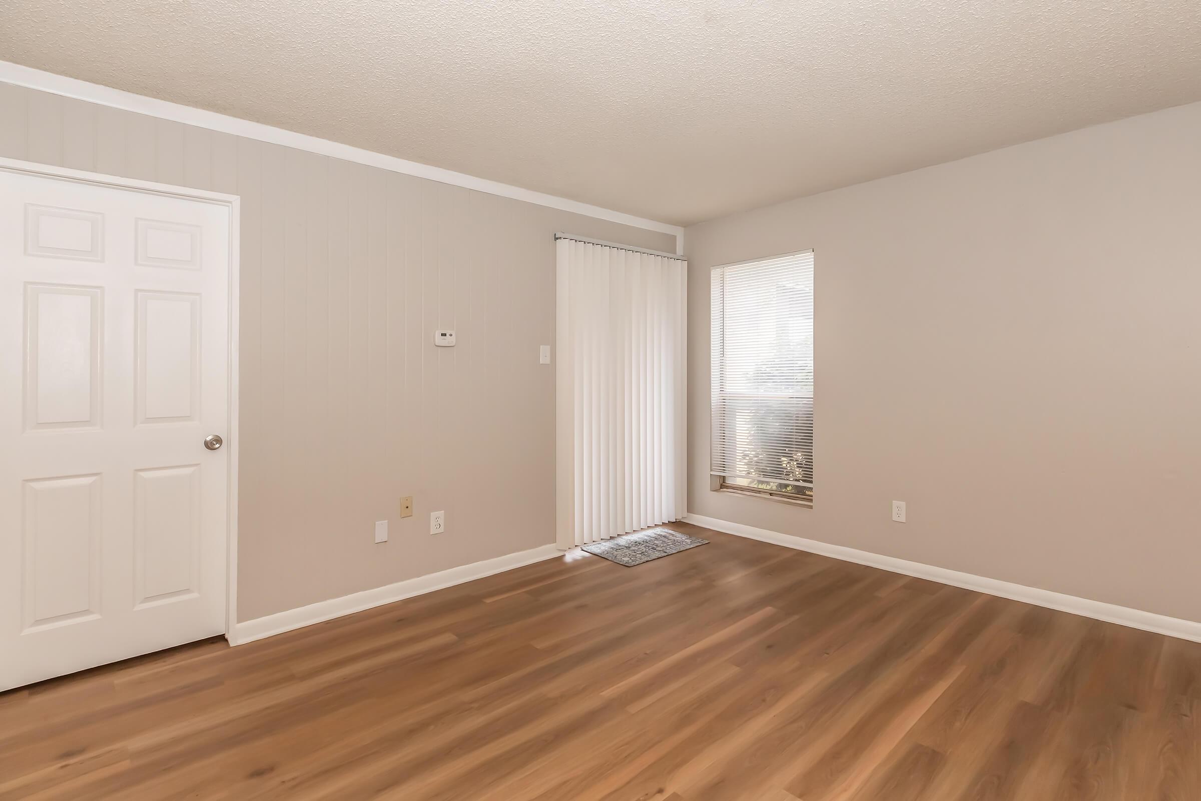 A spacious, empty room with light-colored walls and a smooth wooden floor. There is a closed white door on the left and a window on the right covered with vertical blinds, allowing soft natural light to enter. The room is well-lit and appears ready for furnishing.