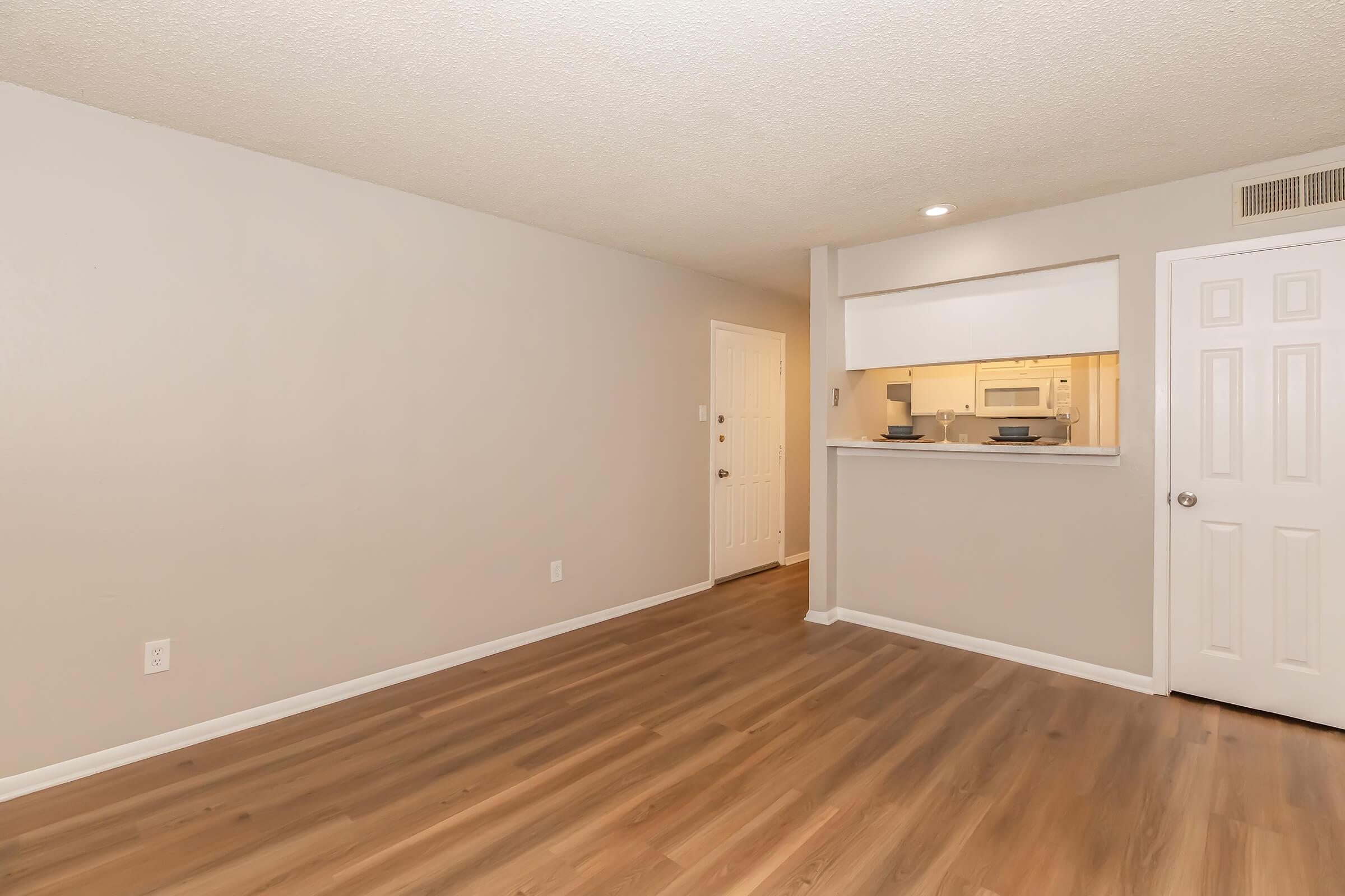 A spacious, empty living room with light-colored walls and a hardwood floor. A doorway leads to a kitchen area, partially visible through an open wall. The entrance door is on the left, and a closed white door is on the right. Soft lighting from recessed ceiling fixtures enhances the bright atmosphere.
