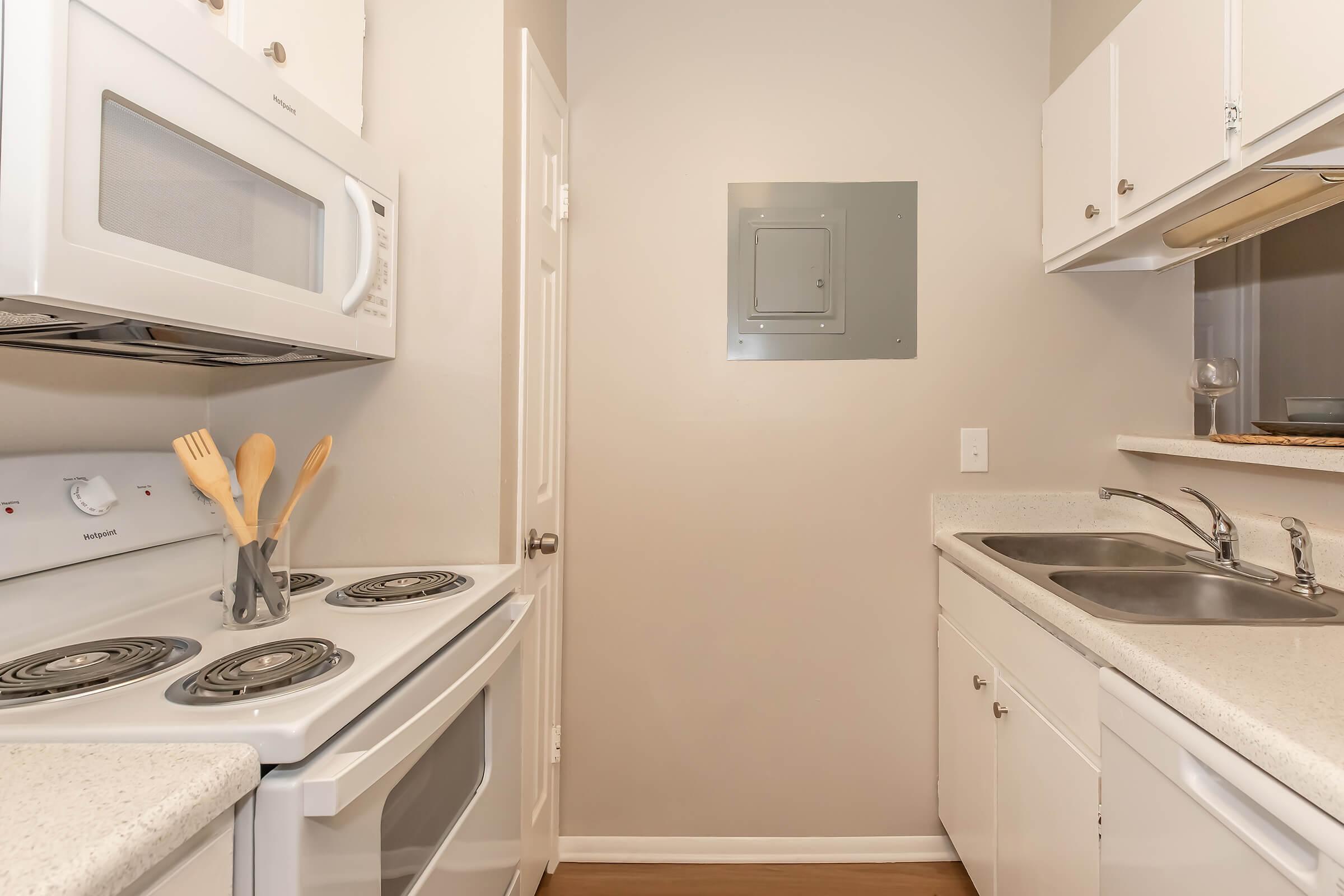 Compact kitchen featuring a white oven, stovetop, and microwave. The cabinetry is light-colored with a clean countertop. A silver vent hood is installed above the sink, and a small access panel is mounted on the wall. The flooring is a light wood finish, creating a bright and functional space.